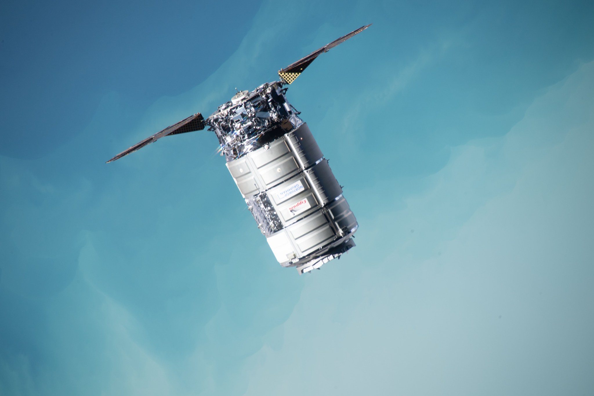 The silver, cylindrical spacecraft is labelled “Cygnus” in red letters and “Northrop Grumman” in blue letters. It has exposed machinery on one end and two solar panels extending like arms on either side of that. In the background is the pale blue Pacific Ocean on Earth below.