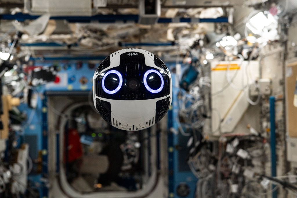 The spherical camera floats in the space station. It is white and black with two large camera lenses on the front that are black with a blue ring around them and look like large eyes and has JAXA in small lettering near the top.