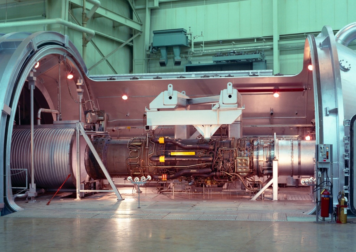 A large engine with many pipes and wires on its sides sits in a large testing facility.