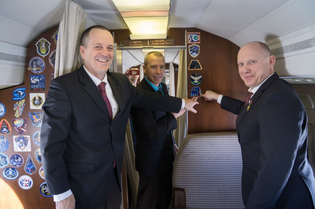 jsc2018e010814 - Aboard a Gagarin Cosmonaut Training Center aircraft en route to their launch site at the Baikonur Cosmodrome in Kazakhstan, Expedition 55 crewmembers Ricky Arnold of NASA (left), Drew Feustel of NASA (center) and Oleg Artemyev of Roscosmos (right) point to their Soyuz crew patch March 4 during their flight from their training base outside of Moscow. The trio will launch March 21 on the Soyuz MS-08 spacecraft for a five month mission on the International Space Station.
