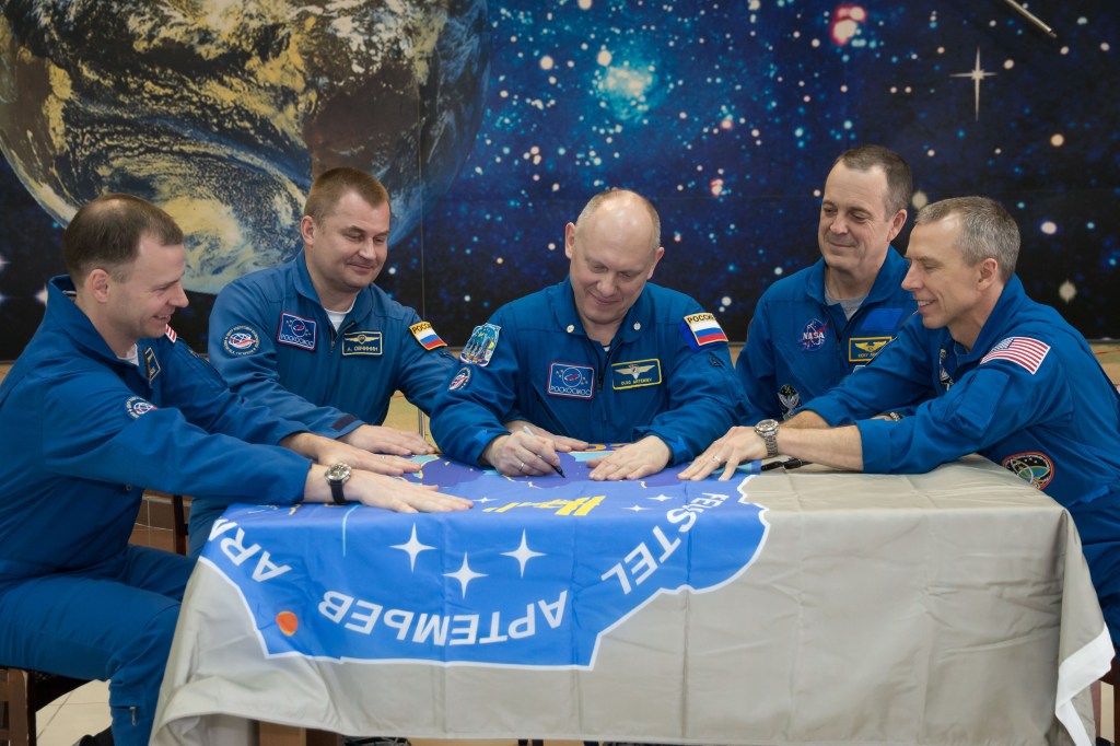 In the Korolev Museum at the Baikonur Cosmodrome in Kazakhstan, the Expedition 55 prime and backup crew members sign a flag bearing their Soyuz MS-08 crew patch insignia March 16 during a traditional preflight tour of the museum. From left to right are backup crewmembers Nick Hague of NASA and Alexey Ovchinin of Roscosmos and prime crewmembers Oleg Artemyev of Roscosmos, Ricky Arnold of NASA and Drew Feustel of NASA, who will launch March 21 on the Soyuz MS-08 spacecraft for a five-month mission on the International Space Station.
