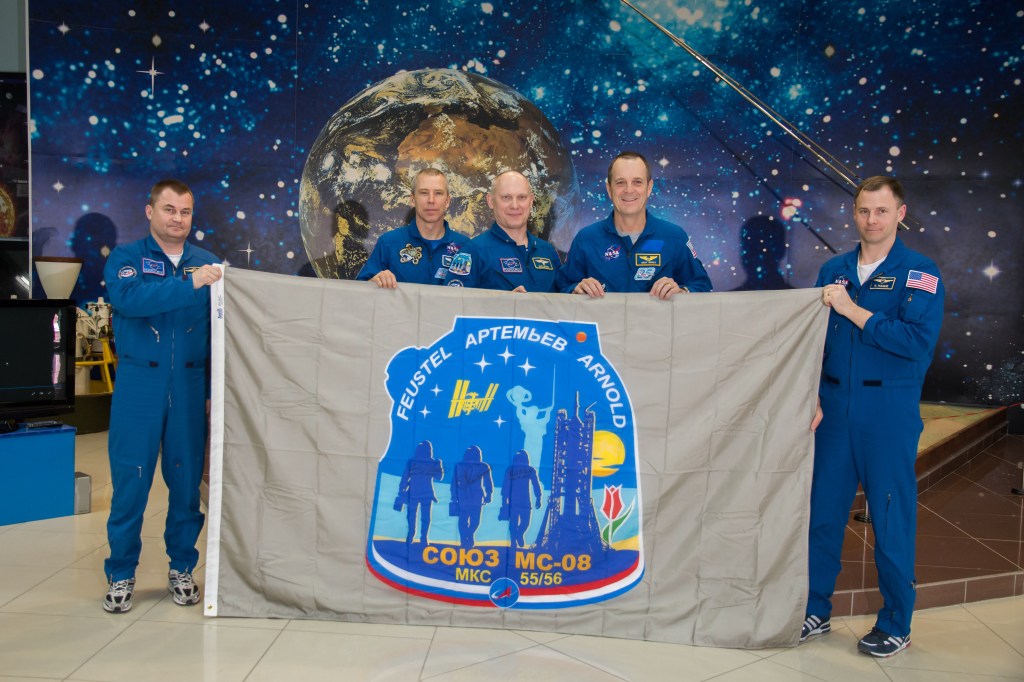 In the Korolev Museum at the Baikonur Cosmodrome in Kazakhstan, the Expedition 55 prime and backup crewmembers display a flag March 16 bearing the Soyuz MS-08 crew patch insignia during a traditional preflight tour of the museum. From left to right are backup crewmember Alexey Ovchinin of Roscosmos, prime crewmembers Drew Feustel of NASA, Oleg Artemyev of Roscosmos and Ricky Arnold of NASA, and backup crewmember Nick Hague of NASA. Feustel, Artemyev and Arnold will launch March 21 on the Soyuz MS-08 spacecraft for a five-month mission on the International Space Station.