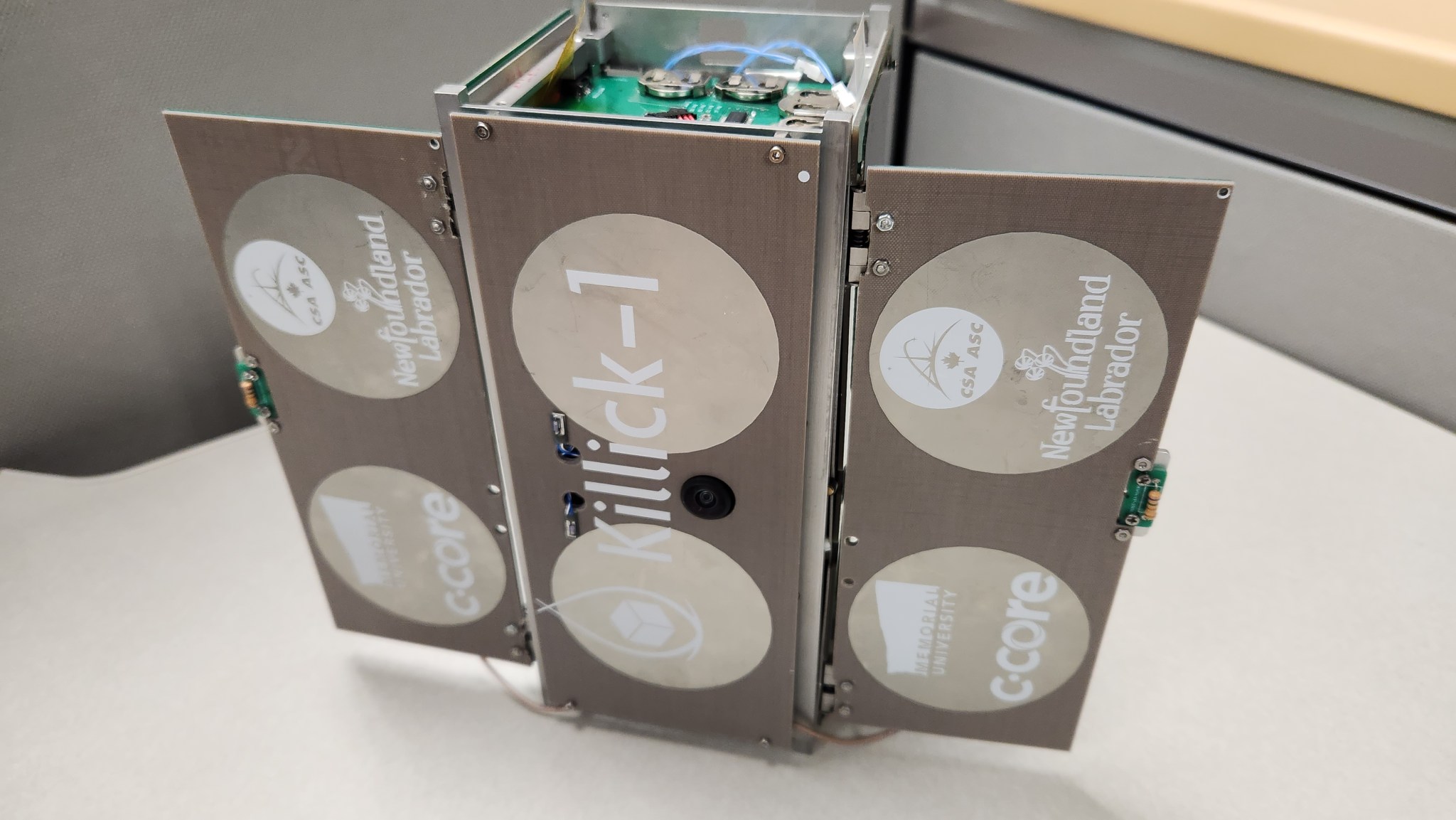 A rectangular box with two sides open stands on its end on a white table. The sides and front have KILLICK-1 and logos for the investigation sponsors on them. A green circuit board with several connected wires is visible on the top of the box.