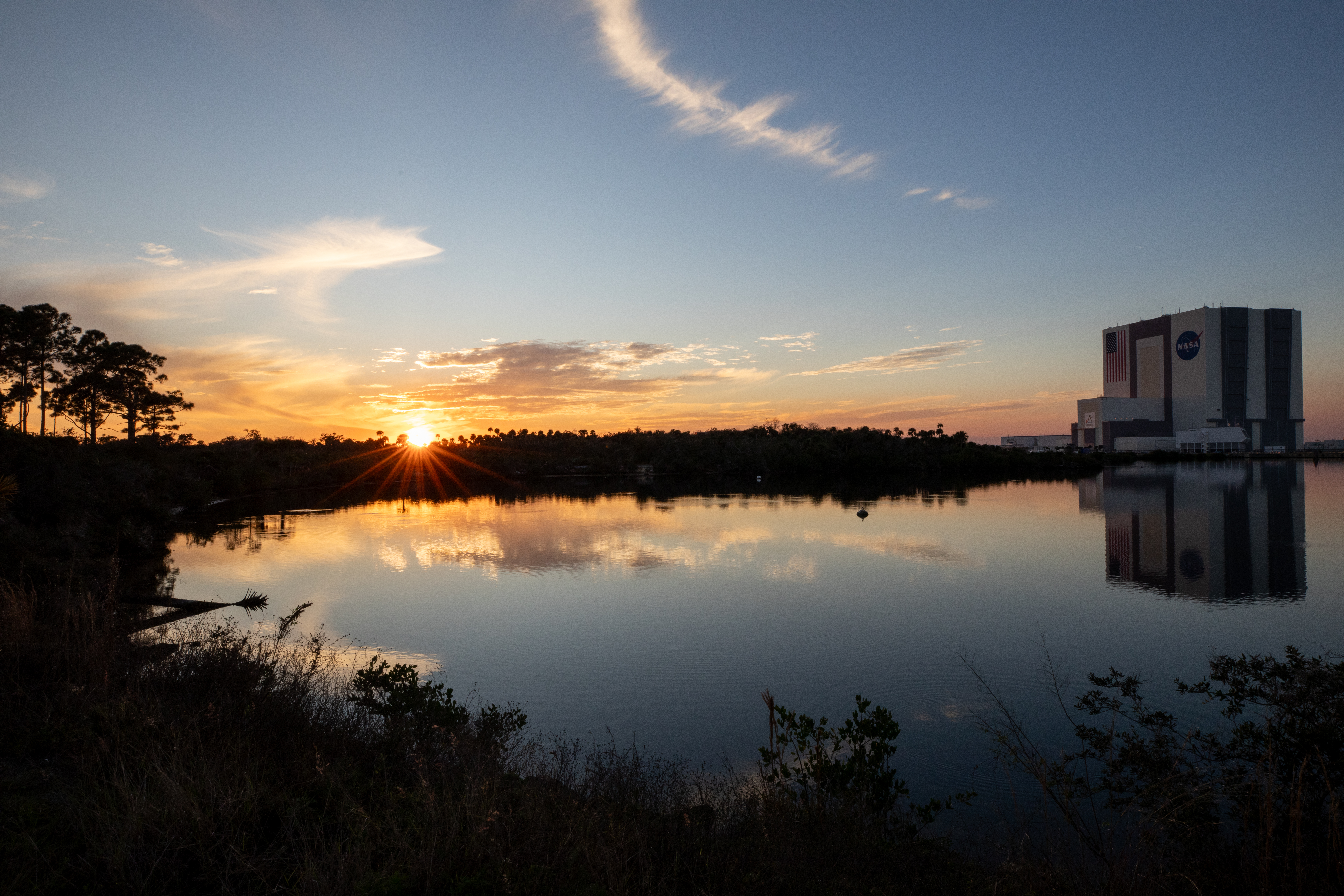 Slightly left of center, the Sun sets behind some vegetation. The sky is a dusty pale blue with a few clouds in it. Trees and plants arc around a body of water, and the Vehicle Assembly Building (VAB) stands at the right edge of the image. The VAB is a very tall, white and gray linear building with a United States flag and NASA meatball logo painted on the front.