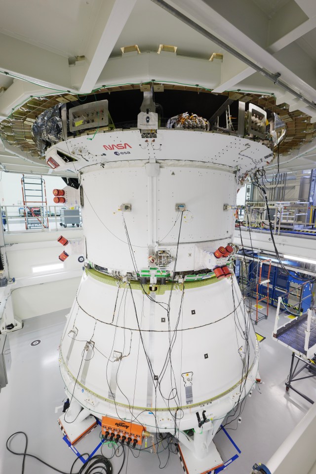 A large white spacecraft component stands in the center of a large room. At the top, the NASA "worm" logo and ESA (European Space Agency) insignia are painted on the crew module adapter.