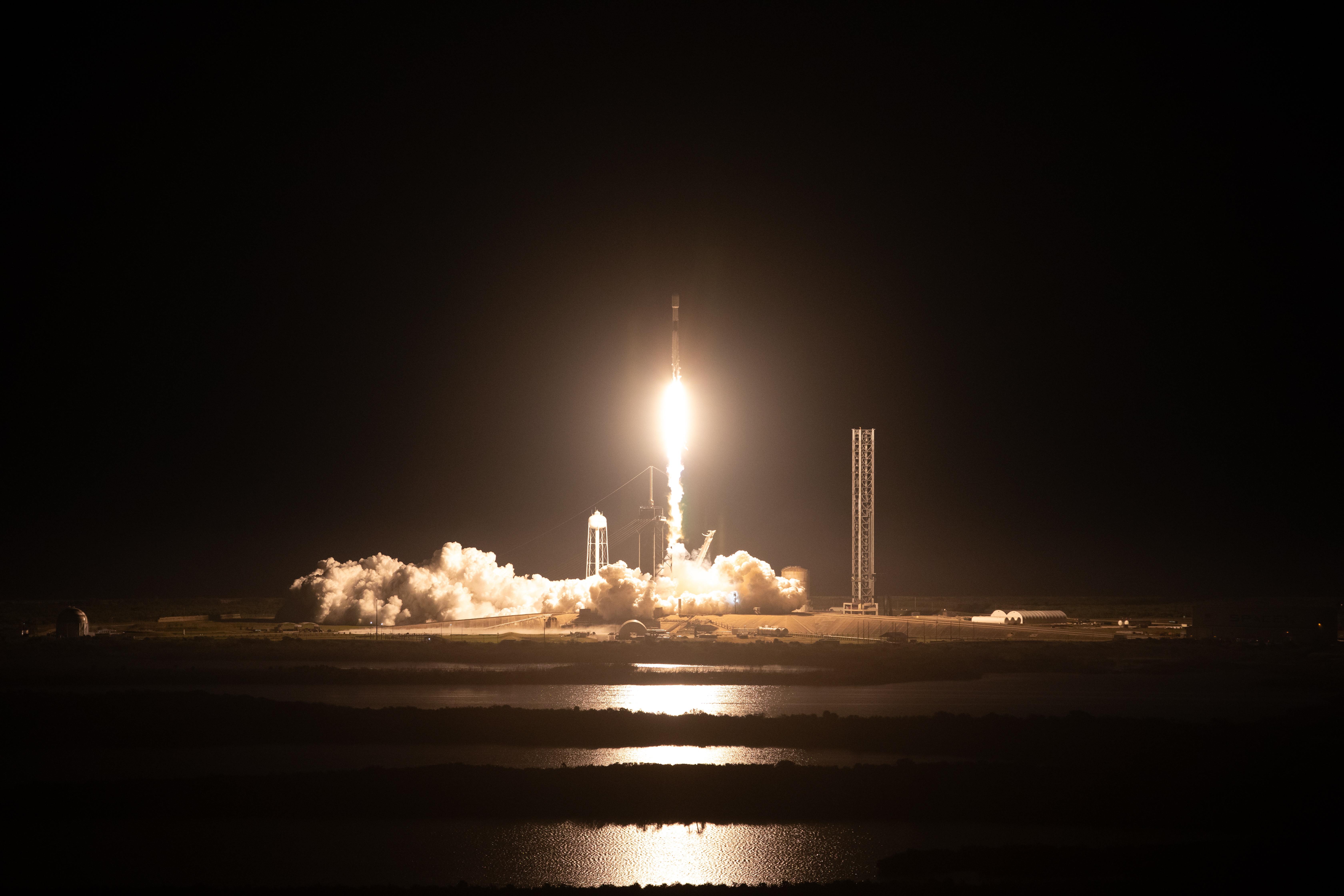 A SpaceX Falcon 9 rocket carrying the Intuitive Machines Nova-C lander takes off from the launch pad at night. The flames coming from the bottom of the rocket (the bright spot at center) light up the surrounding area, illuminating clouds of white vapor that spread outward along the ground. The light also reflects off water in the foreground. Credit: NASA/Kim Shiflett