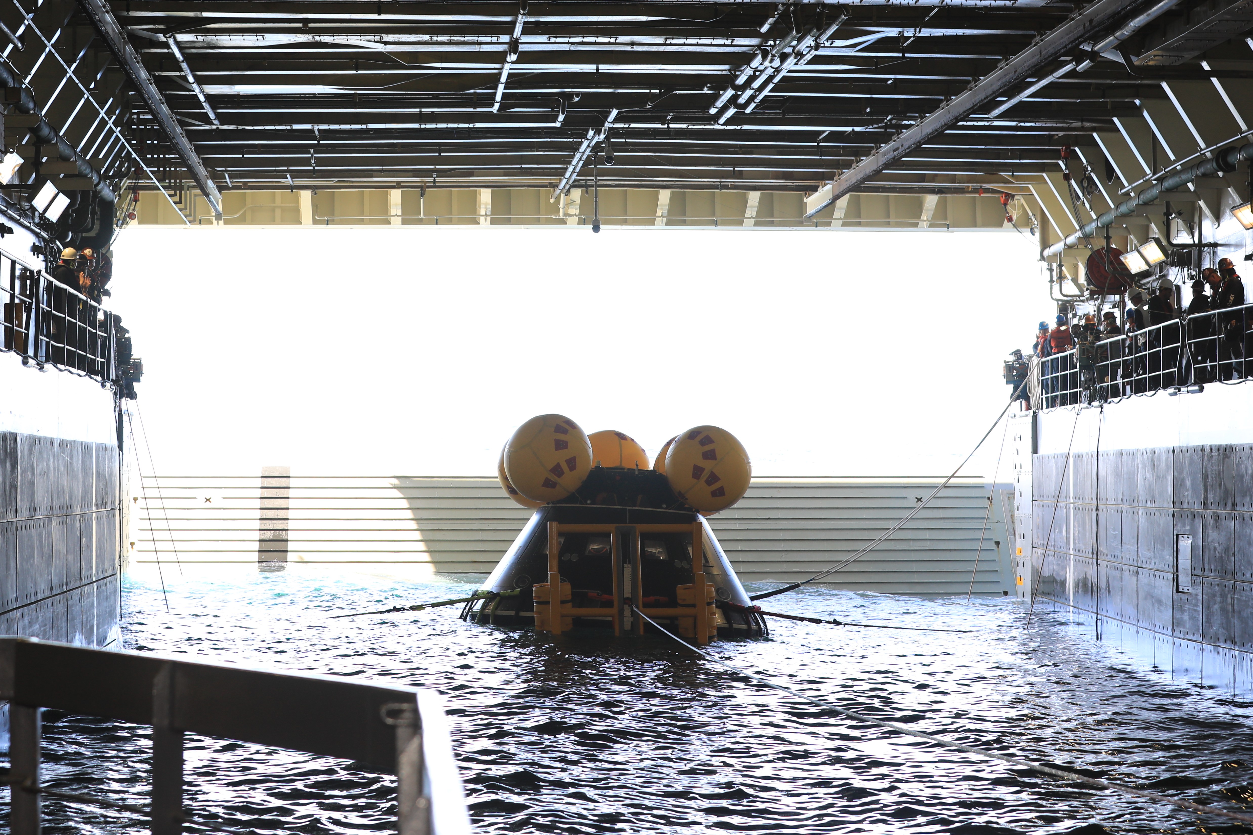 Teams work to secure a test version of NASA's Orion spacecraft on its stand inside a ship's well deck. The well deck has water in it. Straps extend from the spacecraft up to the balconies on either side, where people stand.