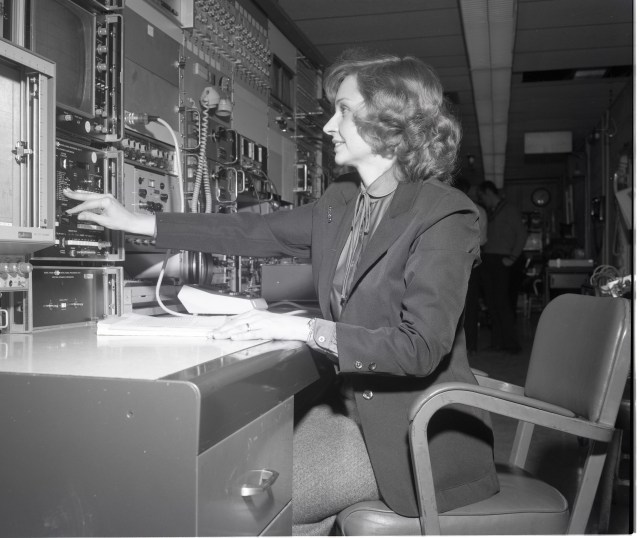 In this black-and-white photo, Loretta Shaw, who is wearing a dark-colored blazer, sits at a desk and reaches forward to adjust a knob on the equipment - filled with switches, buttons, and cords - in front of her.
