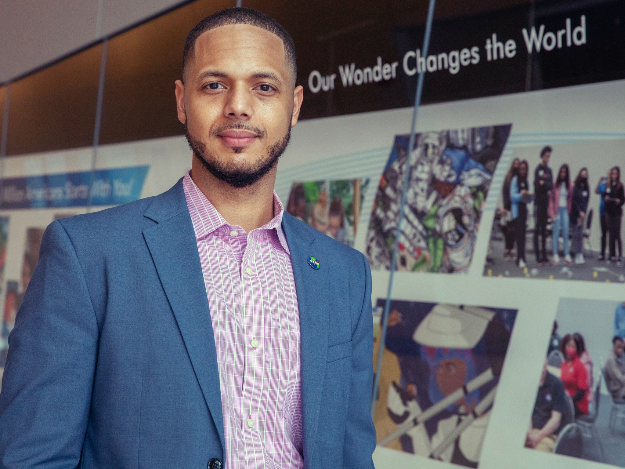 This is a photo of Matthew Hayes, a DEIA Project Analyst with NASA’s Langley Research Center. Matthew is wearing a blue suit and is standing in front of a mural featuring photos of Langley employees. There is text above the photo collage that reads "Our Wonder Changes the World."