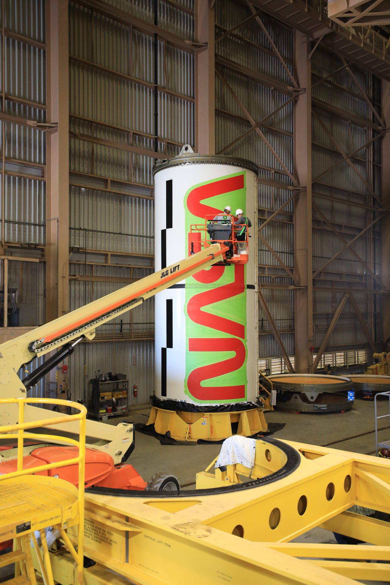 A red NASA "worm" logo is being painted by a person in a bucket event on a solid rocket booster segment inside a building 