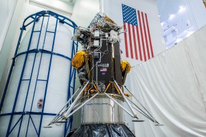 The Intuitive Machines Nova-C lander for the company's first Commercial Lunar Payload Services delivery is positioned before being encapsulated inside its launch fairing. The lander is yellow, grey, black, and white and an American flag and NASA and Intuitive Machines logos can be seen on the spacecraft’s surface. The background of the facility that the spacecraft is in is white, and a large American flag can be seen on the wall behind it.