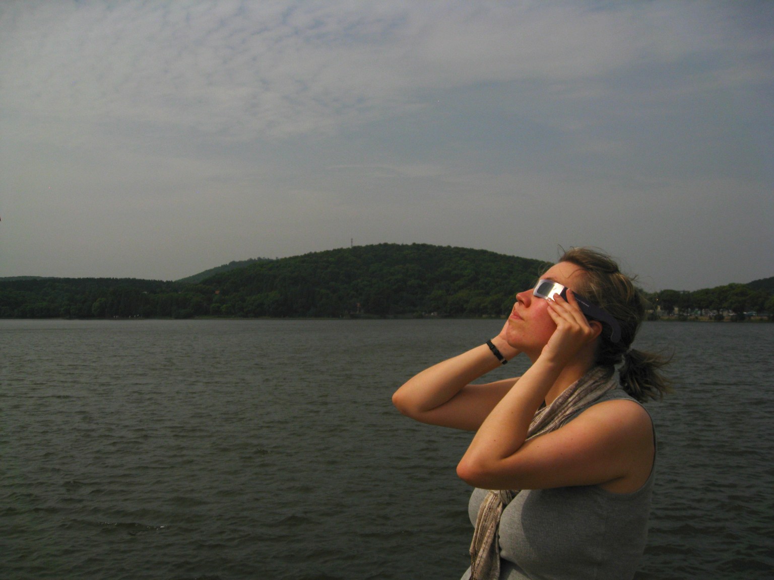 A woman stands in front of a lake, looking up, wearing eclipse glasses.