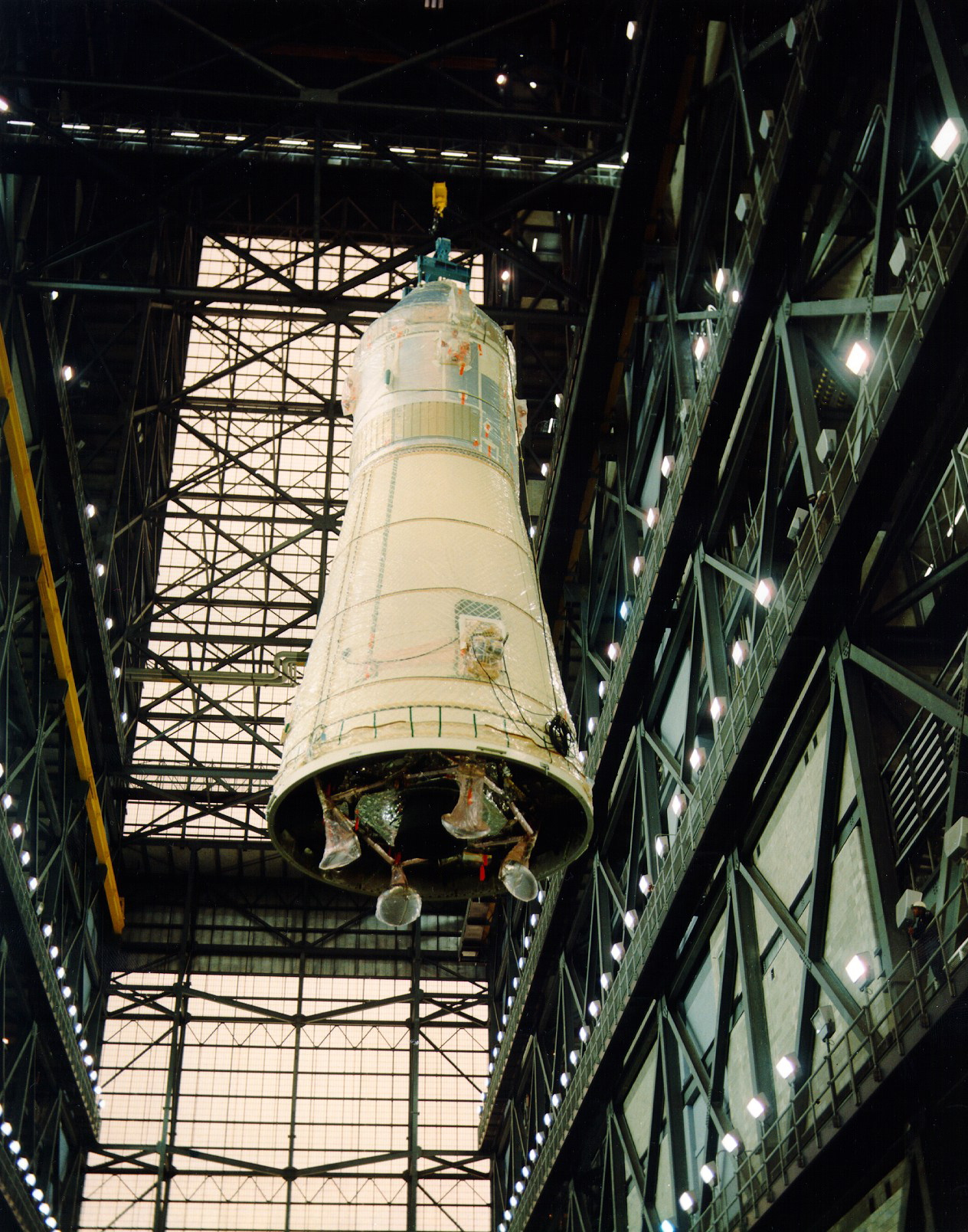 Workers lift the spacecraft for stacking onto the rocket, the footpads of the LM’s folded landing gear visible