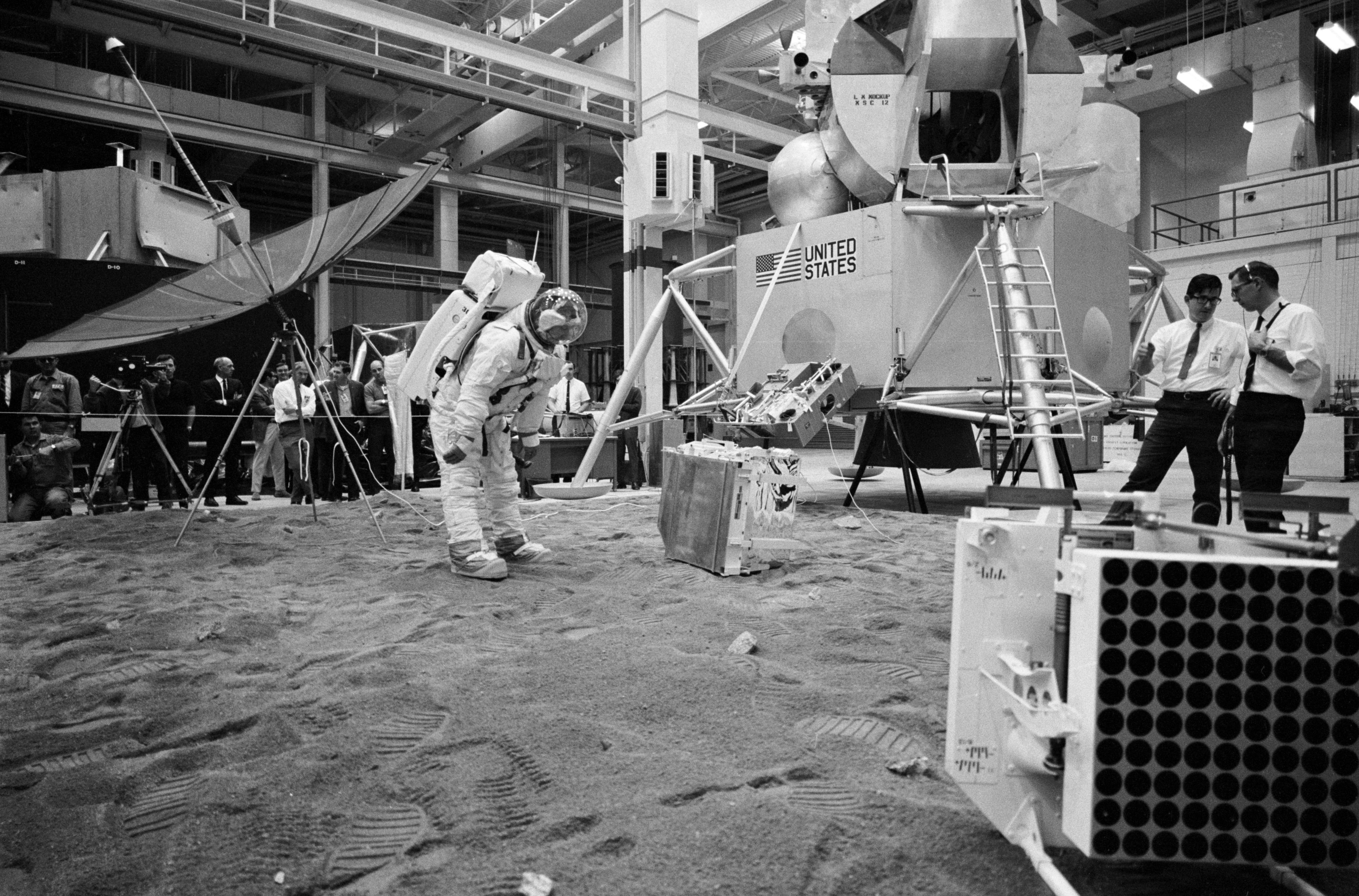 Astronaut Don L. Lind, suited, practices deploying the EASEP instruments as Aldrin, in white shirt behind the dish antenna, oberves