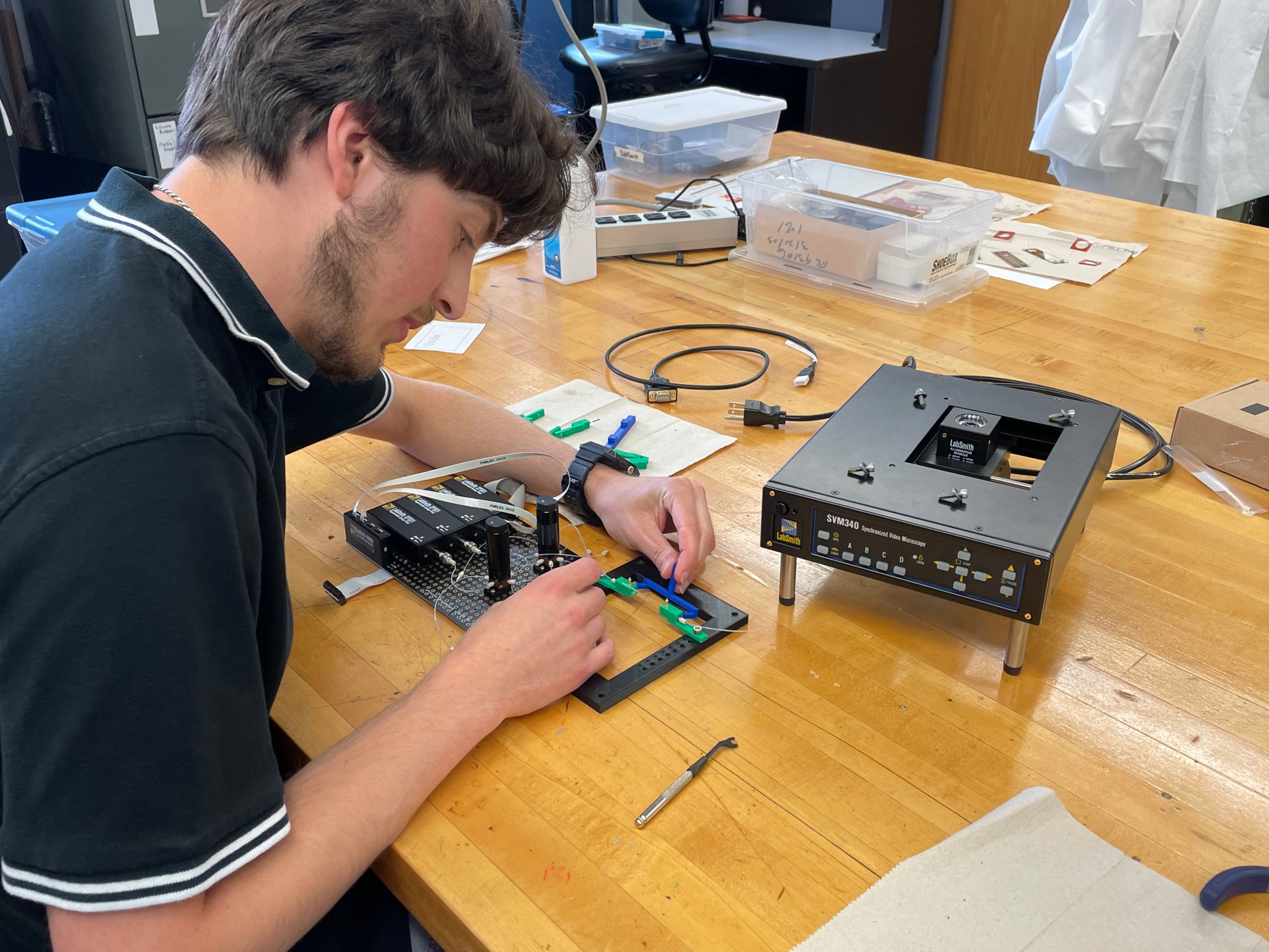 A young man wearing a black short-sleeved polo leans over a wooden workbench, using both hands to attach wires to a small black breadboard. A black rectangular box sits on the bench next to it.