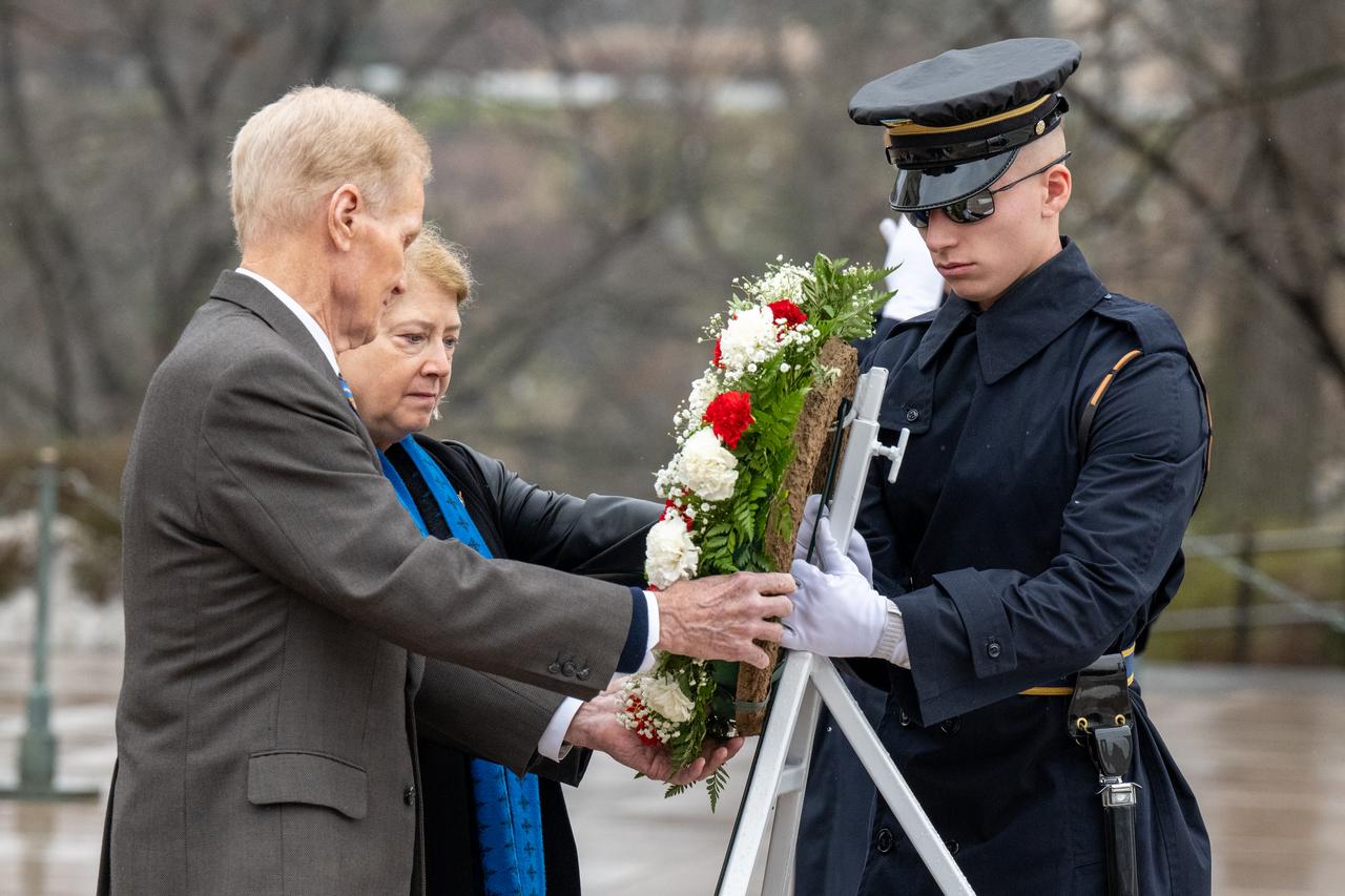 NASA Administrator Bill Nelson and NASA Deputy Administrator Pam Melroy lay a wreath of red and white carnations on a white stand. A soldier assists them by holding the stand for the wreath.