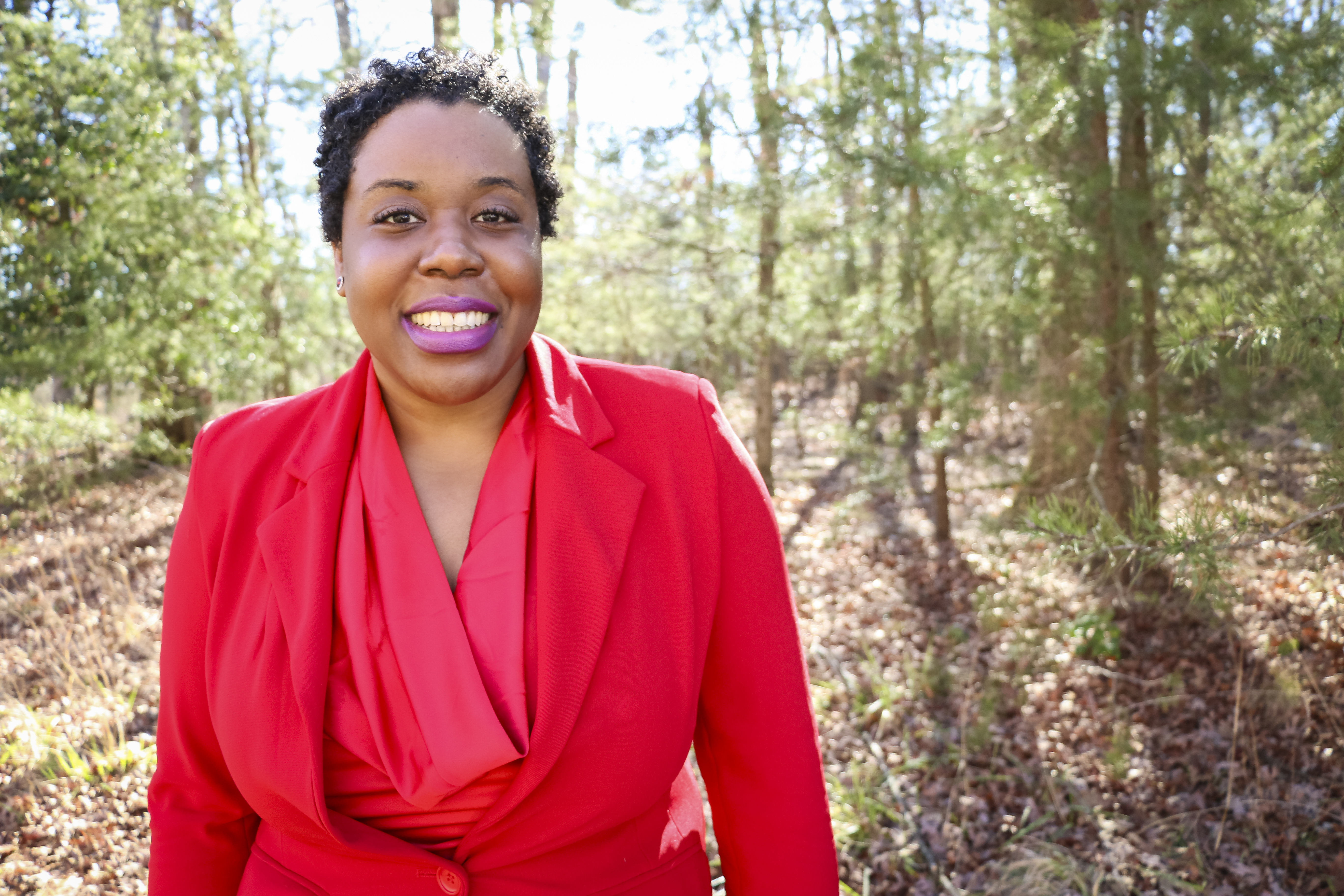 A woman that's standing in the woods wearing a red blouse and red suit jacket smiles widely at the camera.