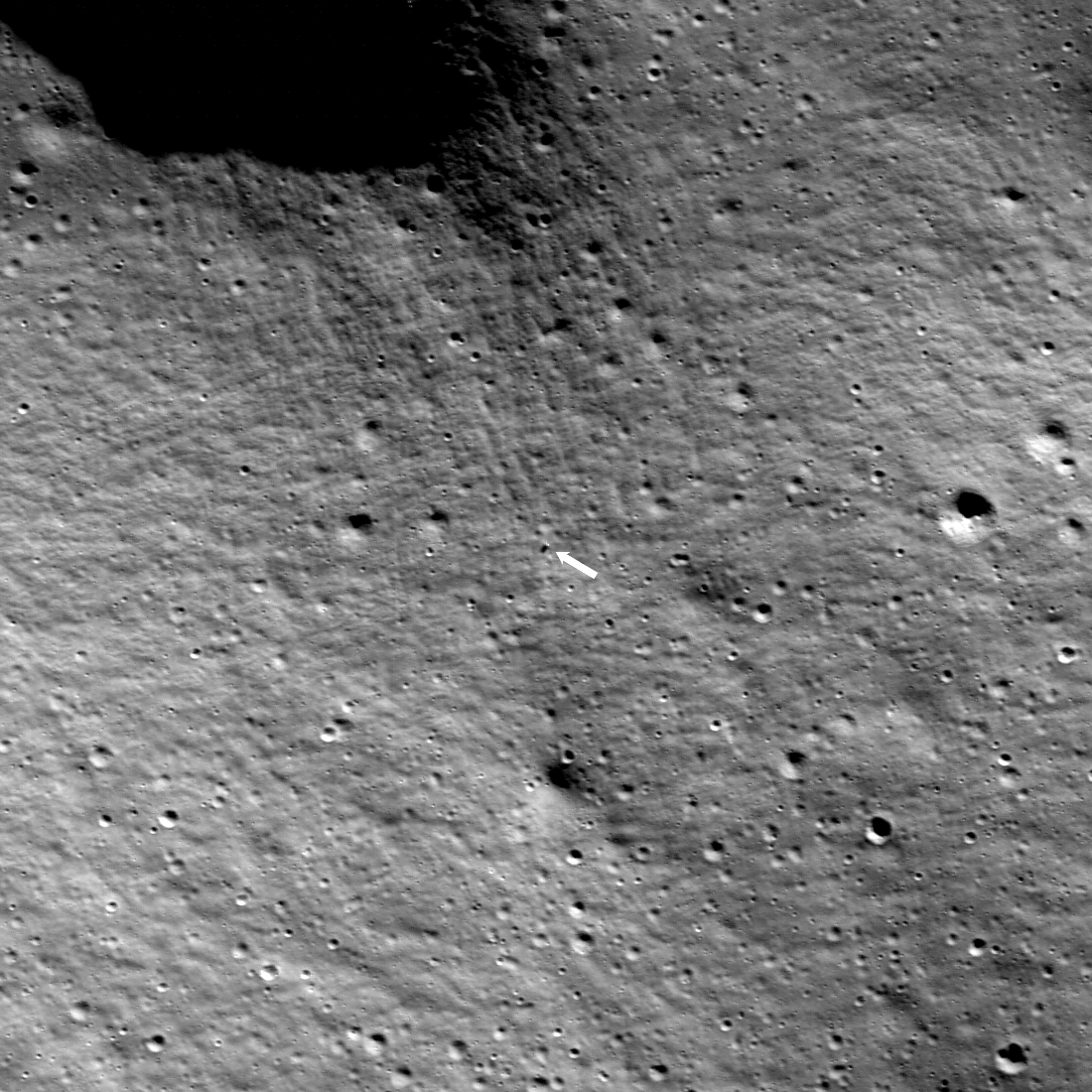 grayscale image of Moon's rocky surface from above, with a small white dot at the center of the frame: IM-1 lander, location indicated with white arrow