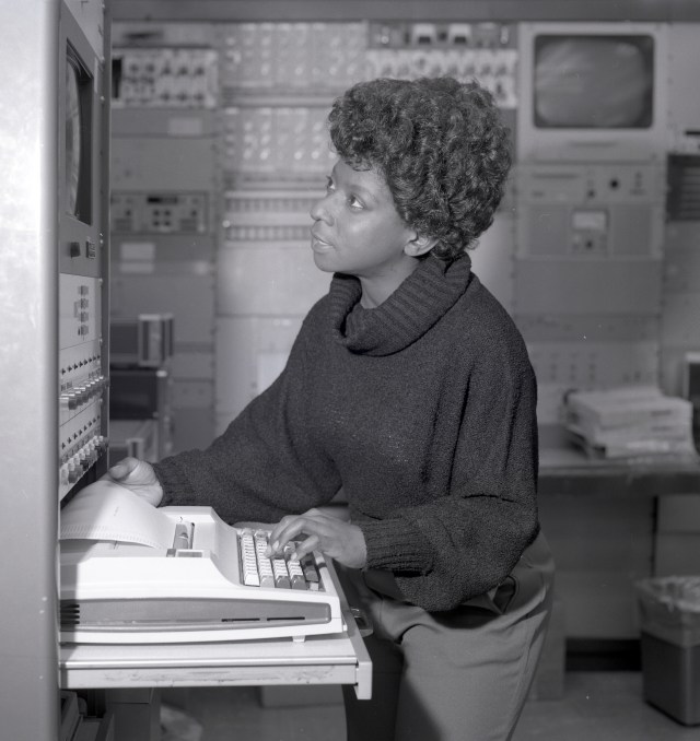 In this black-and-white photo, Phyllis Williamson, who is wearing a dark-colored sweater, types on a keyboard that's connected to a machine as she looks up at the screen above it. Behind her are other pieces of computer equipment and screens.