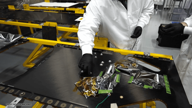 two people in white suits and black gloves work with dark-hued electronics on a table