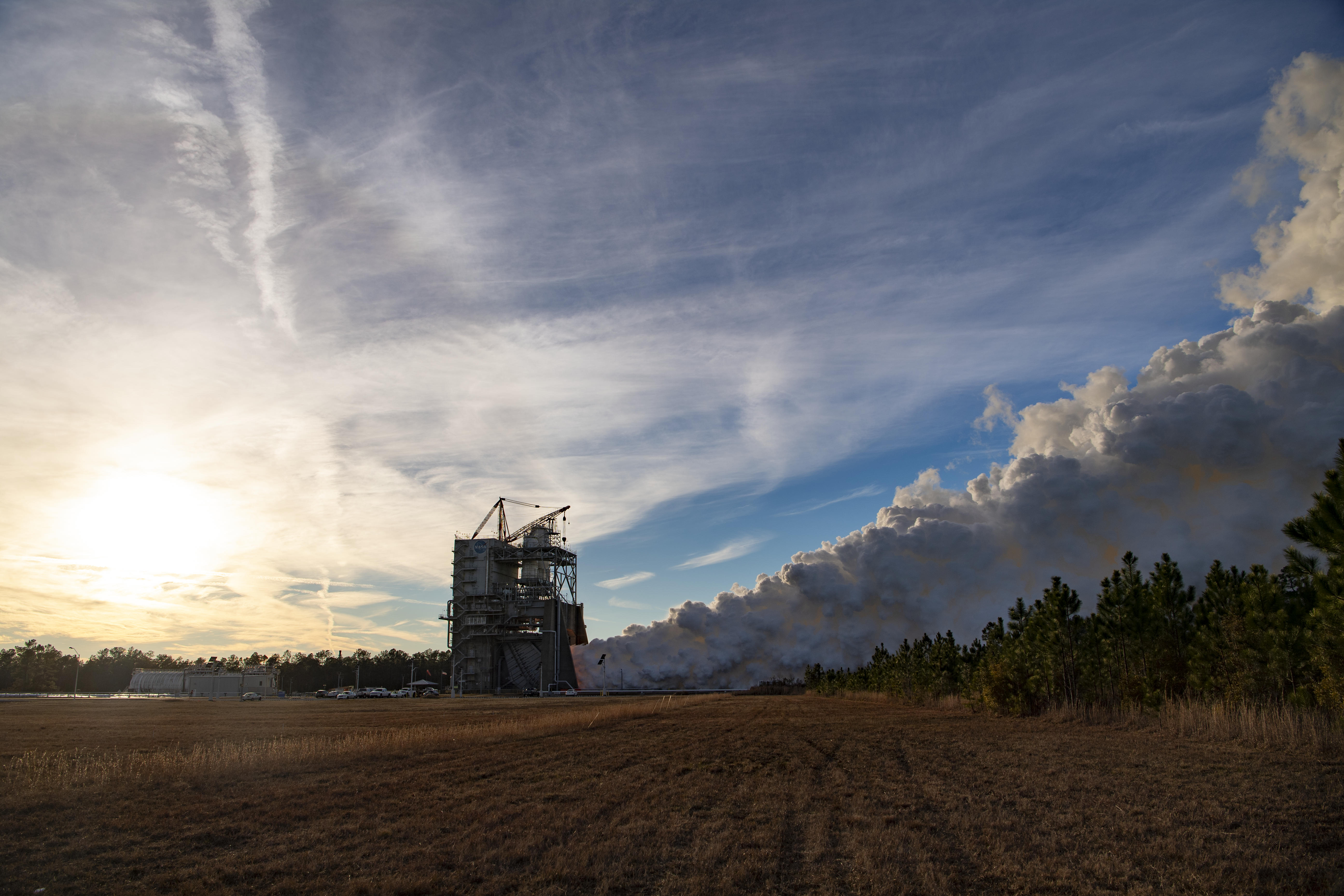 White vapor clouds billow up and to the right of this image during a hot fire test. The sun is a diffuse, bright yellow spot, covered by wispy clouds that dominate the sky.