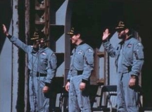 From aboard the U.S.S. New Orleans, Skylab 4 astronauts Gerald P. Carr, left, Edward G. Gibson, and William R. Pogue wave to the crowd assembled dockside at North Island Naval Air Station (NAS) in San Diego