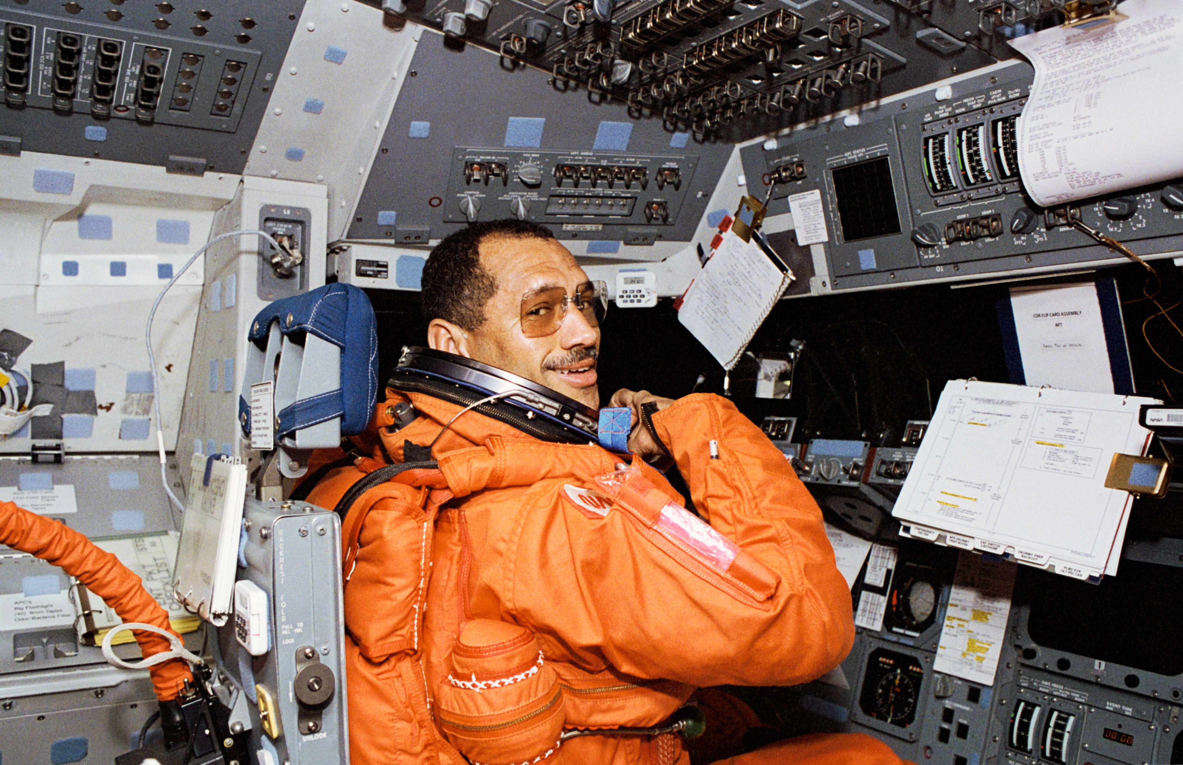 Astronaut Charles F. Bolden, a Black man, looks over his right shoulder and smiles at the camera. He is wearing an orange launch and entry suit and square tinted glasses without temples. He is sitting at the commander's station, which has many switches and dials, along with a notebook, a 3-ring binder, and various sheets of paper.
