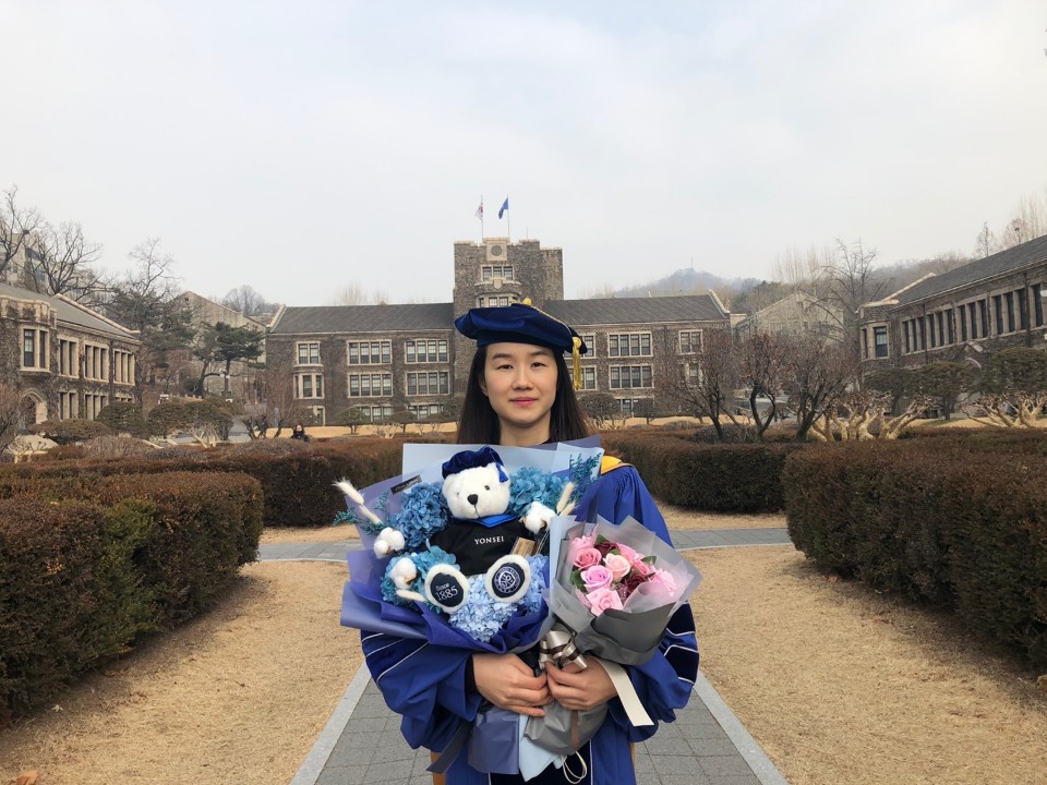 Sujung Go stands in front of a courtyard and building at Yonsei University in South Korea wearing a graduation cap and gown, holding flowers and a stuffed bear
