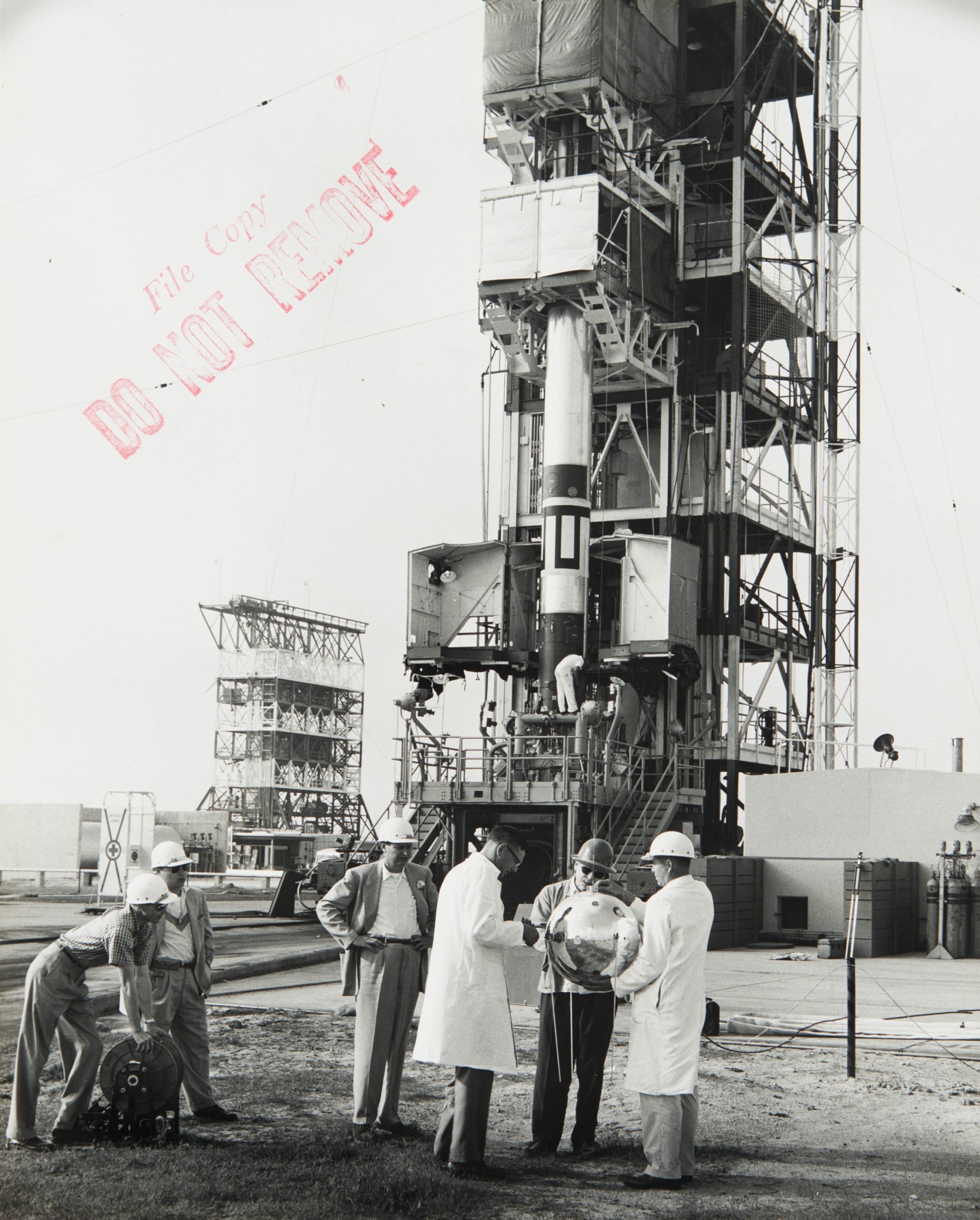 Men hold the Vanguard 2 satellite in the foreground with the Vanguard SL-V poised for launch in the background