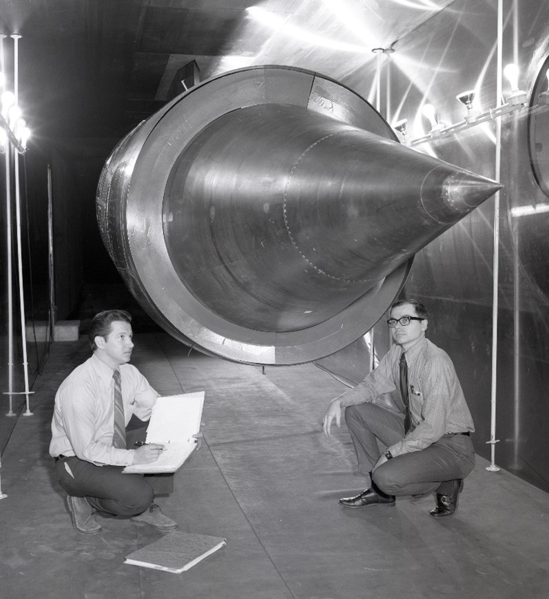 A black-and-white photo of two people dressed in suits and ties crouching under a YF-12 flight inlet in a wind tunnel. The person on the left looks up at the large, pointy inlet and writes in a binder.