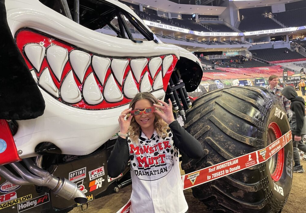 A woman wearing a shirt with the words, Monster Mutt Dalmatian, poses by a huge truck with big wheels while holding solar eclipse viewing glasses in front of her eyes.