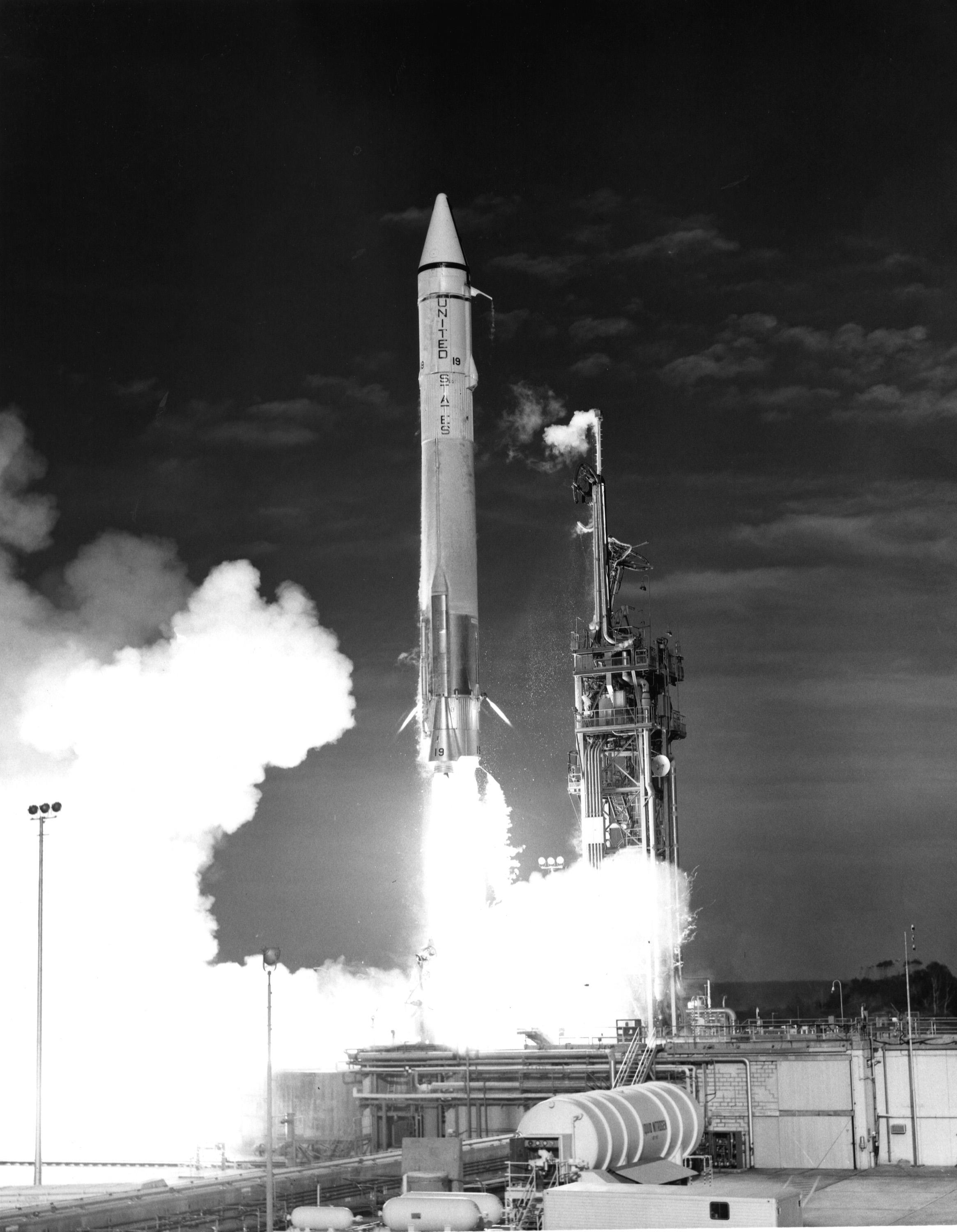 A black-and-white photo of the Mariner 7 launch. An Atlas-Centaur rocket takes off, flames pouring out underneath it and smoke billowing off to the left. The words "United States" are written on the rocket. A few wispy clouds are visible in the sky.