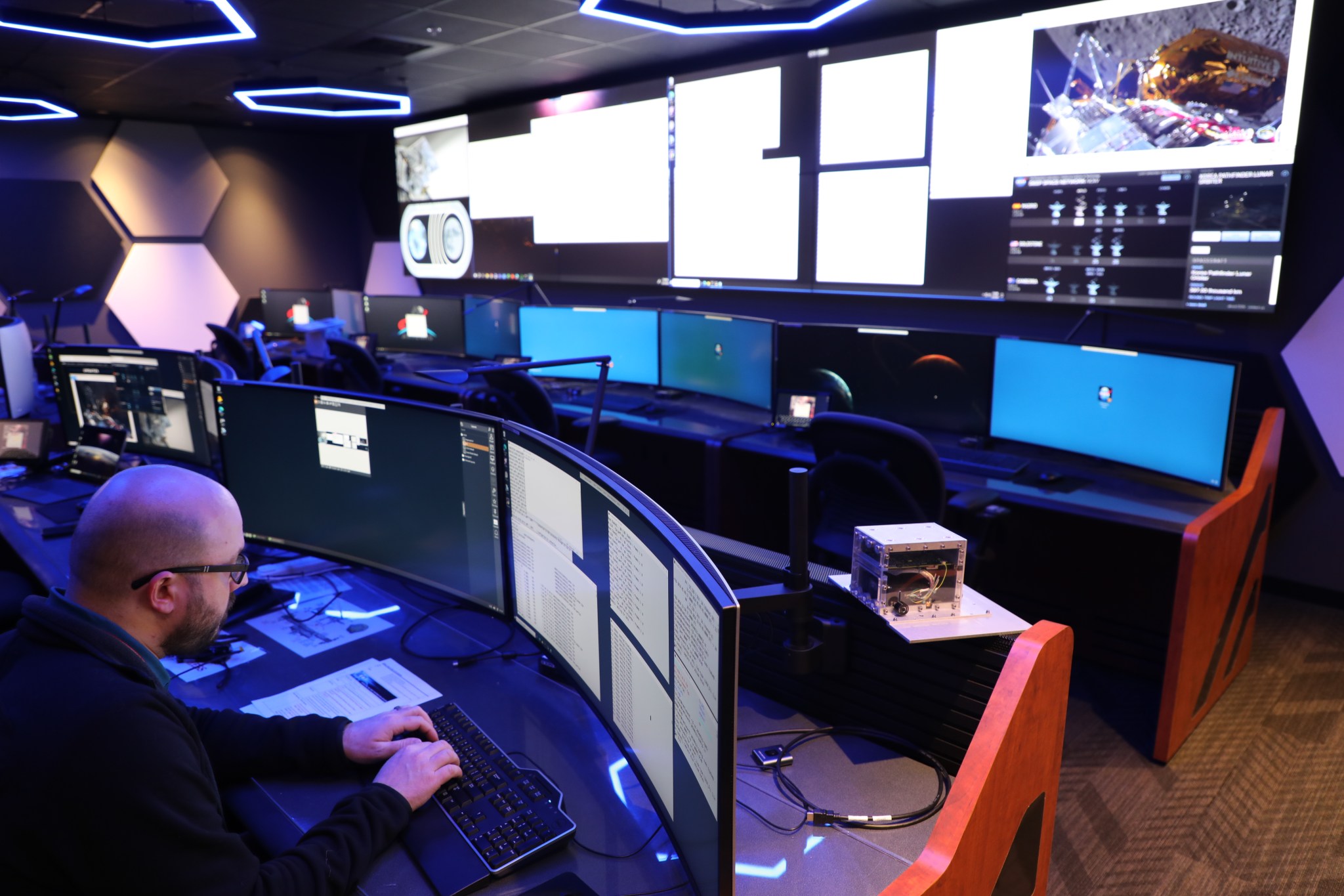 A man sits in front of a computer screen in a large control room with huge screens in the background.