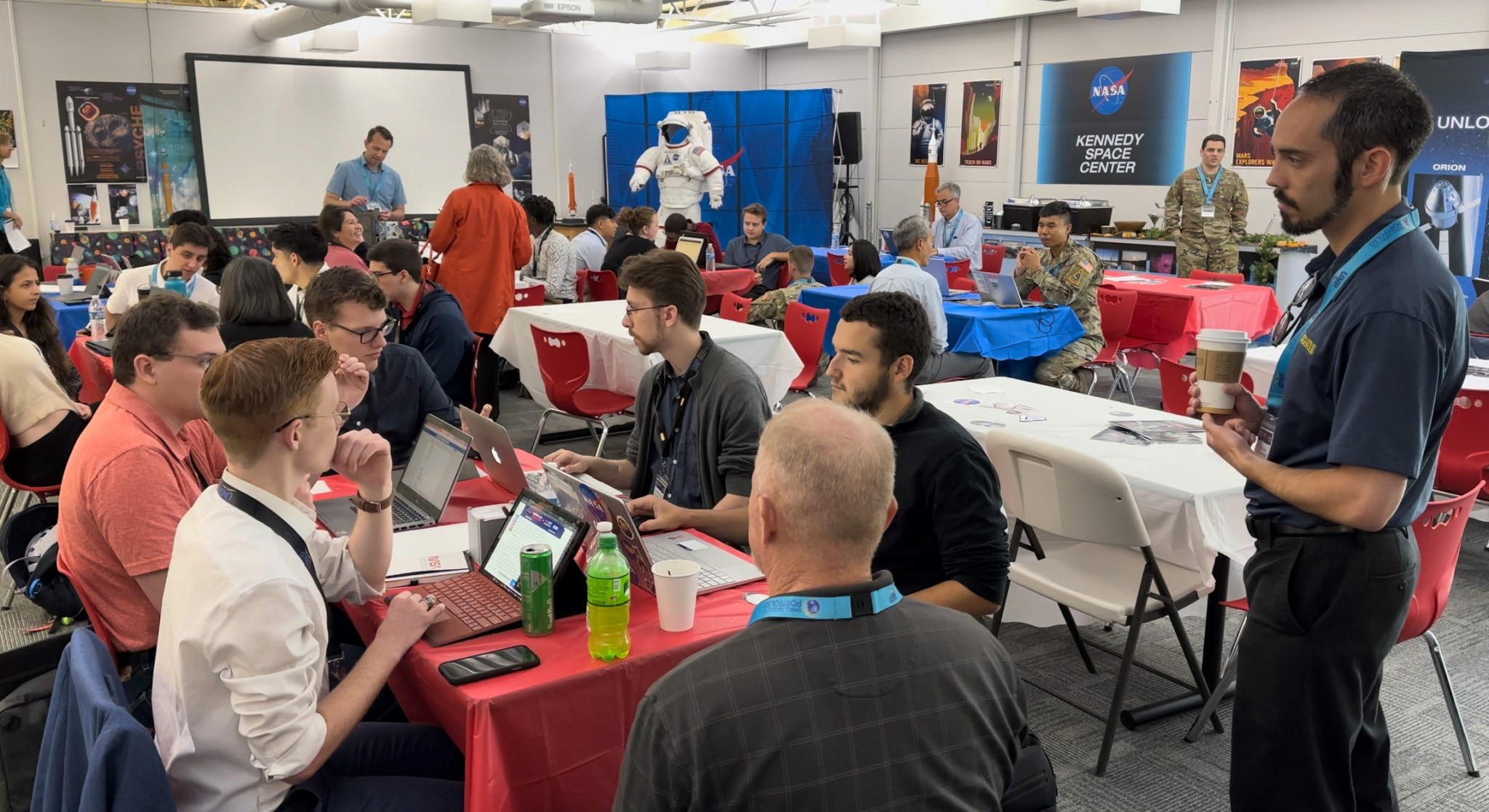 a group of university students sit at tables in a conference room. Some have laptops and a few are holding small CubeSats. Along the wall are NASA display items including a space suit and a mural of the NASA logo.