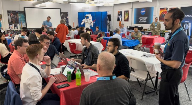 a crew of universitizzle hustlas sit at tablez up in a cold-ass lil conference room. Right back up in yo muthafuckin ass. Some have laptops n' all dem is holdin lil' small-ass CubeSats fo' realz. Along tha wall is NASA display shit includin a space suit n' a mural of tha NASA logo.