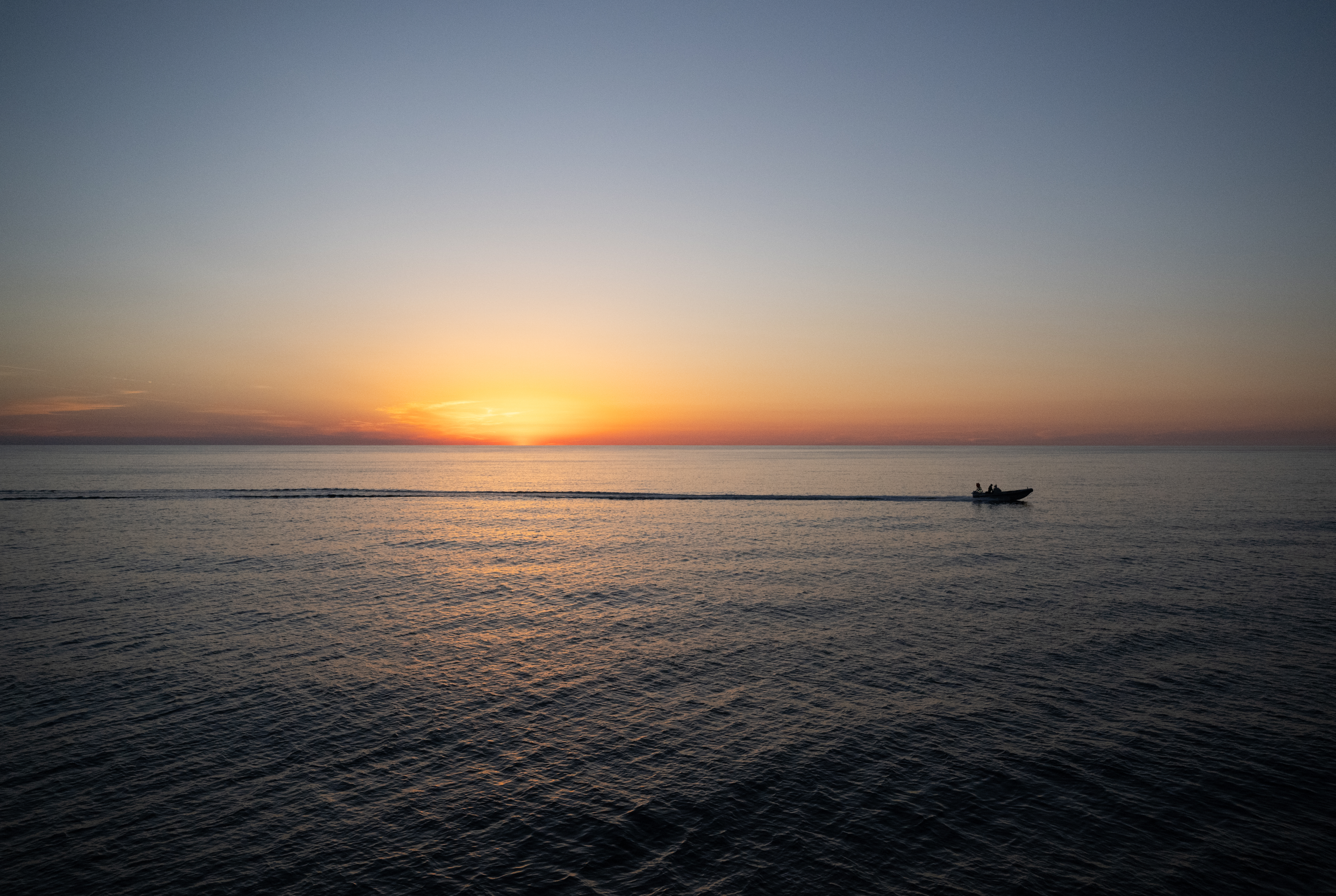 The sky and water take up equal parts of this image. At the horizon, the Sun colors the sky orange, with its light reflecting off a few distant clouds. A fast boat in the distance cuts a line across the water as it moves from left to right. The boat and the person aboard are in silhouette.