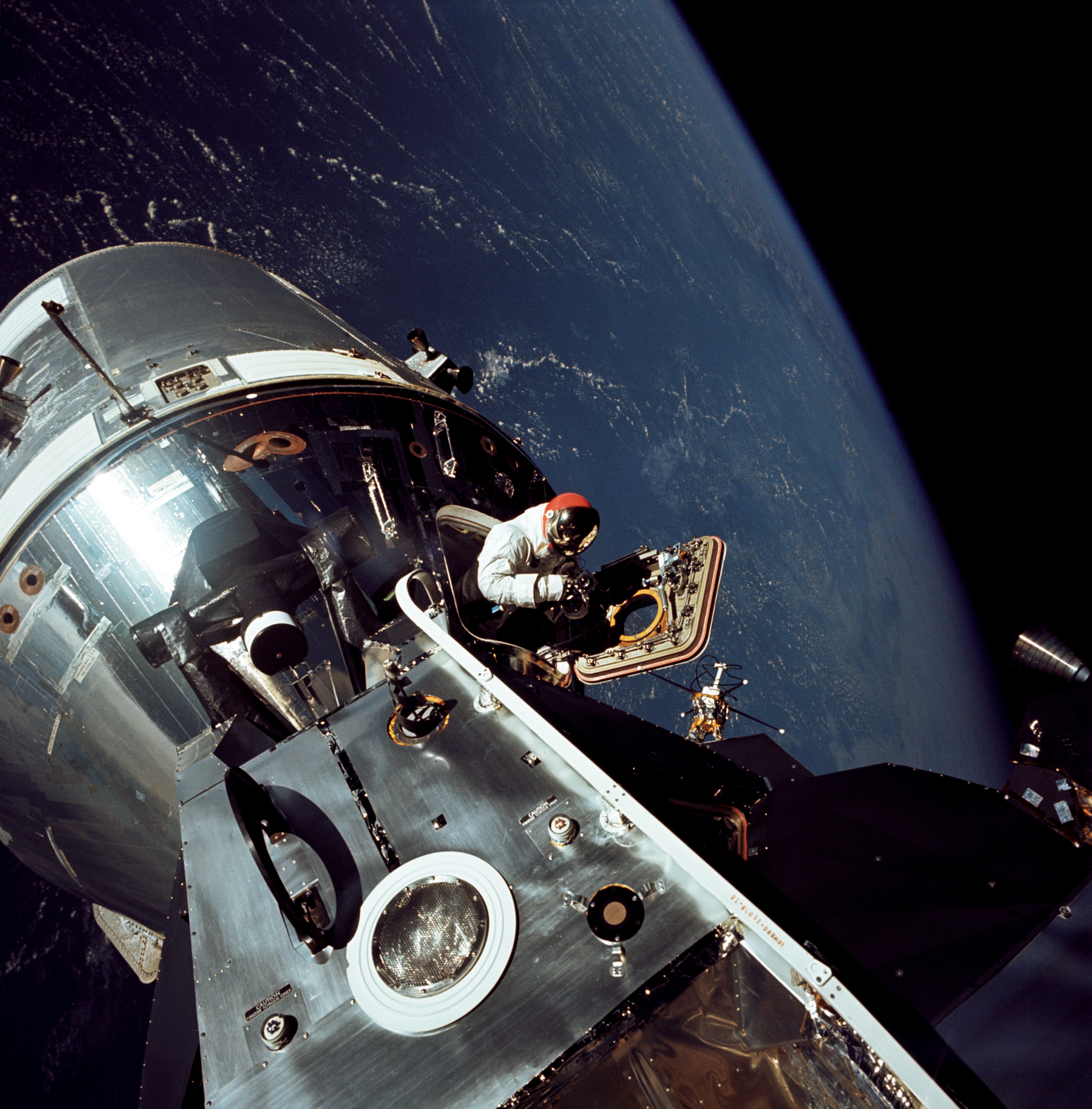 Earth is a deep blue backdrop to this photo of astronaut David Scott and the command module and lunar module of the Apollo 9 mission. The joined modules are a metallic silver. He stands in the open hatch of the command module while wearing a white spacesuit. A metallic visor tops his red helmet.