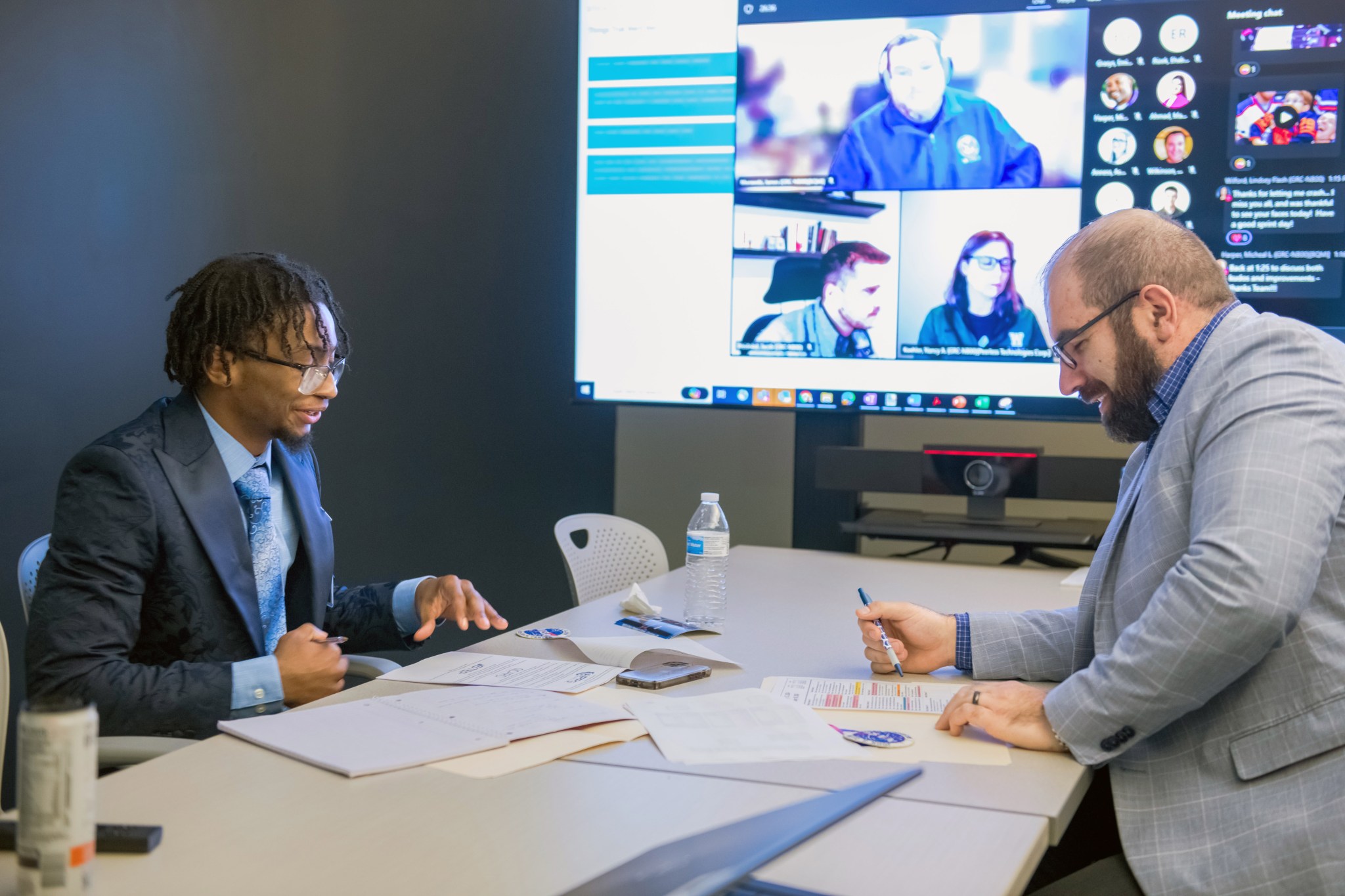 A well-dressed college student, left, and a subject matter expert sit across from each other at a large desk. A large screen with faces of online meeting attendees shows on a large screen in the background.