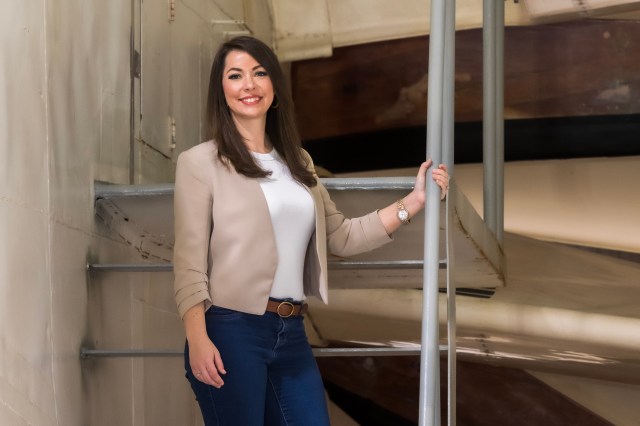 A woman with long brown hair and dark brown eyes stands against metal stairs, smiling. She holds a railing with her left arm and her right arm down at her side. She's wearing a light beige blazer over a white shirt, blue jeans, and a brown belt.
