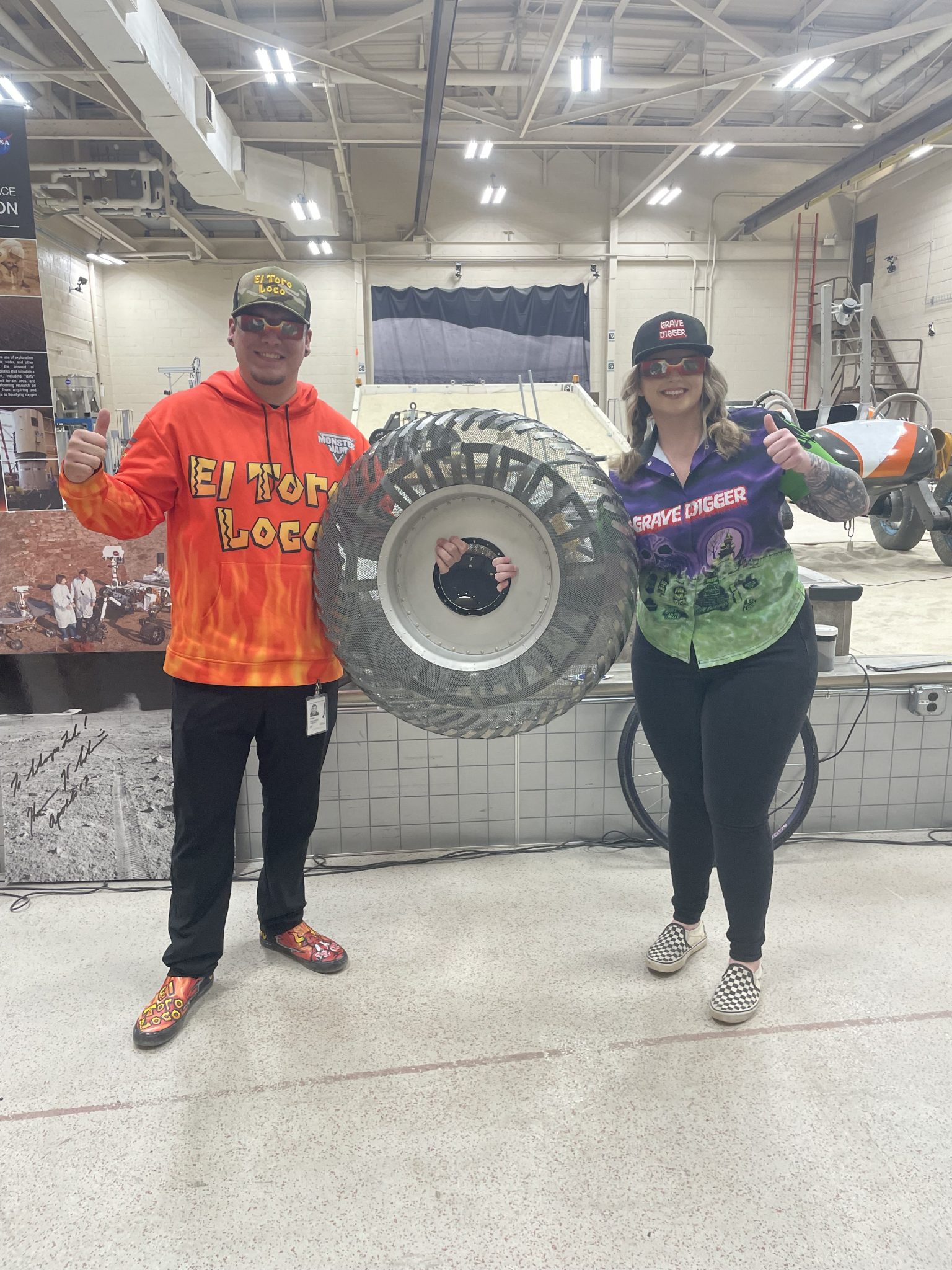 Colorfully clothed Monster Jam drivers hold a lunar tire between them in front of a brightly lit large sandbox-type testing area.