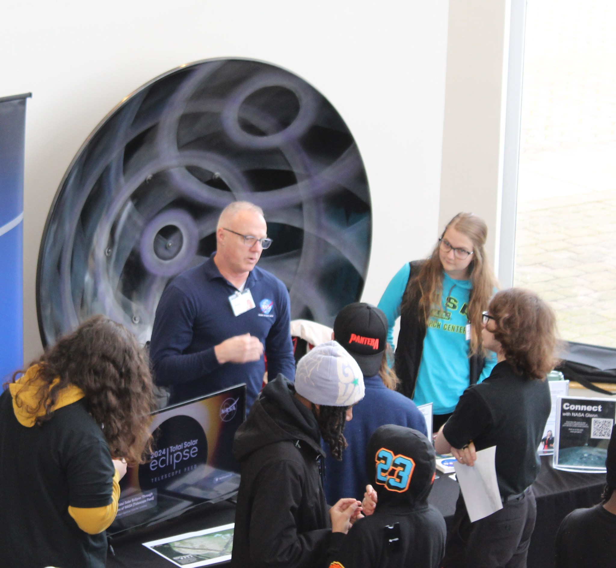 Two representatives from NASA stand in front of a table surrounded by students who are asking questions. Information on the eclipse and NASA Connect is on the table.