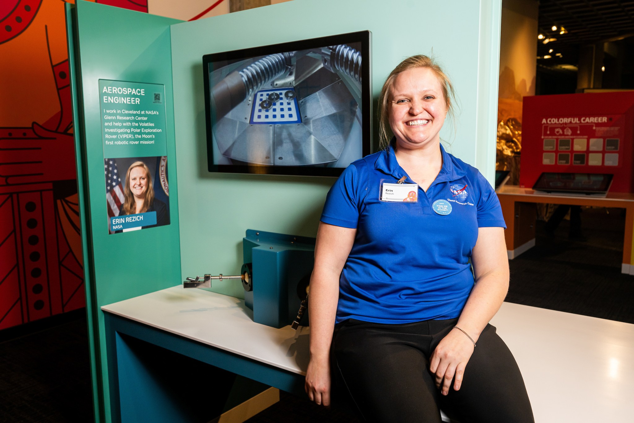 A NASA employee sits on the edge of a table with hardware behind her. A video of her work is on a screen, and her photo and credentials are displayed.