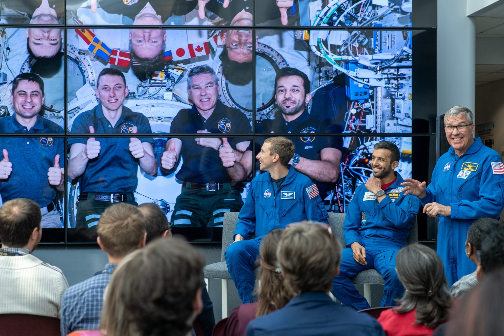 Three astronauts in blue flight suits stand in front of a bank of TV monitors