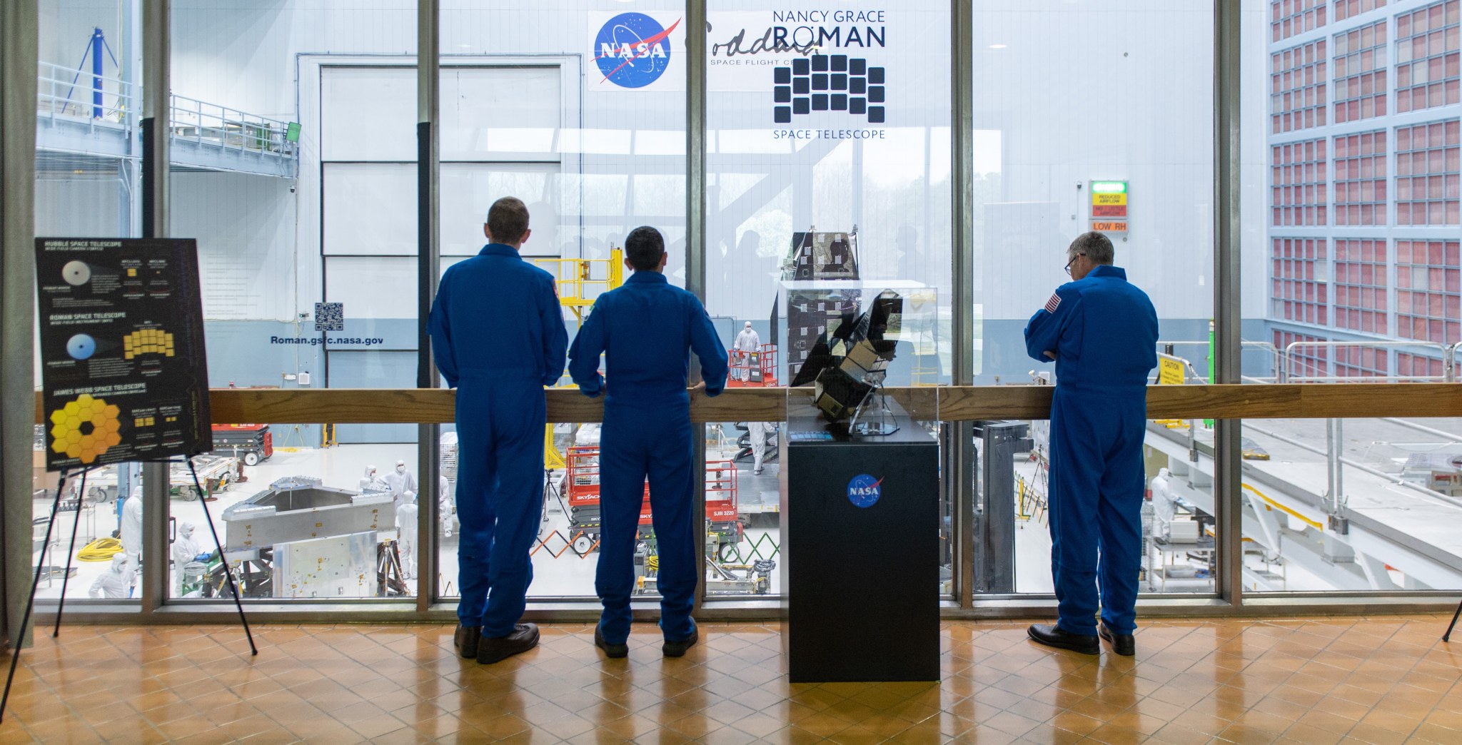 with backs to the camera, three astronauts in blue flight suits stand before glass windowpanes that look in on the enormous clean room where spaceflight hardware for the Roman Space Telescope can be seen in the background