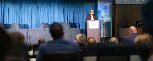 far shot of Dr. Makenzie Lystrup speaking at a podium against a curtained backdrop; attendees in foreground