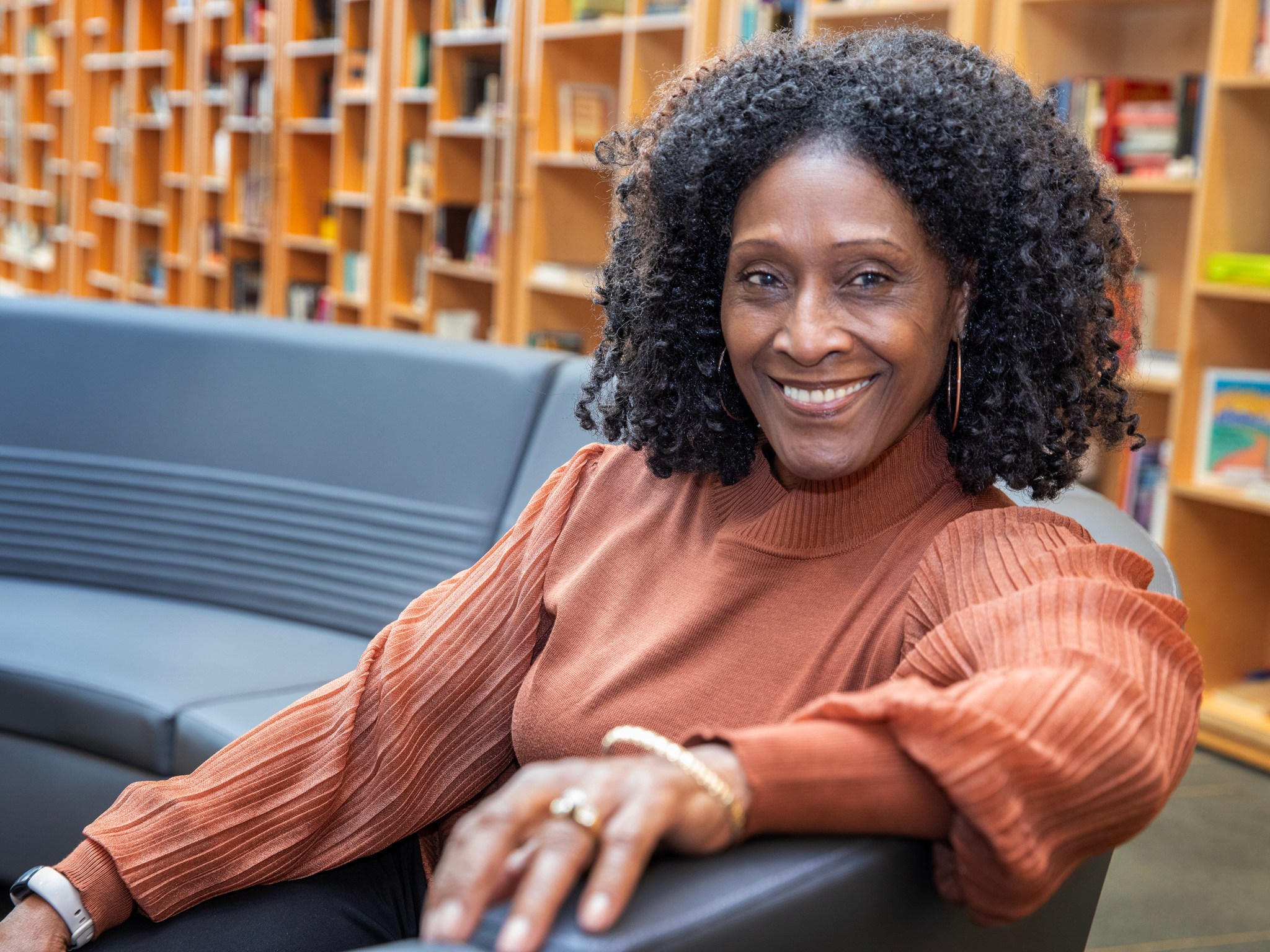 Gwendolyn Wheatle is an Administrative Assistant in the Office of STEM Engagement at NASA Langley Research Center. She is pictured in this photo smiling and wearing an orange blouse. Gwendolyn is sitting on a couch, and bookshelves can be seen in the background of the photo