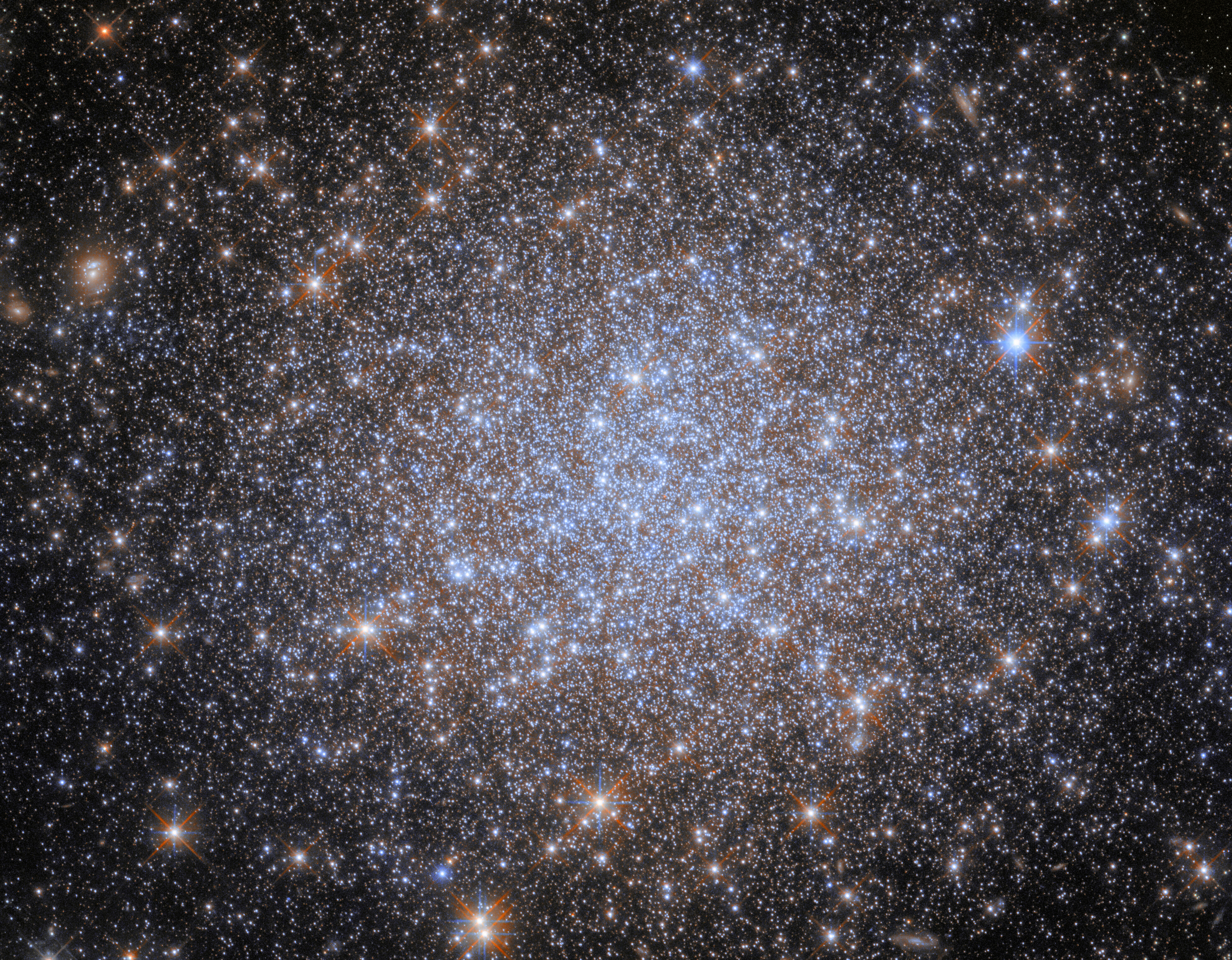 A cluster of stars. Most of the stars are very small and uniform in size, and they are notably bluish and cluster more densely together toward the center of the image. Some appear larger in the foreground. The stars give away to a dark background at the corners of the image.