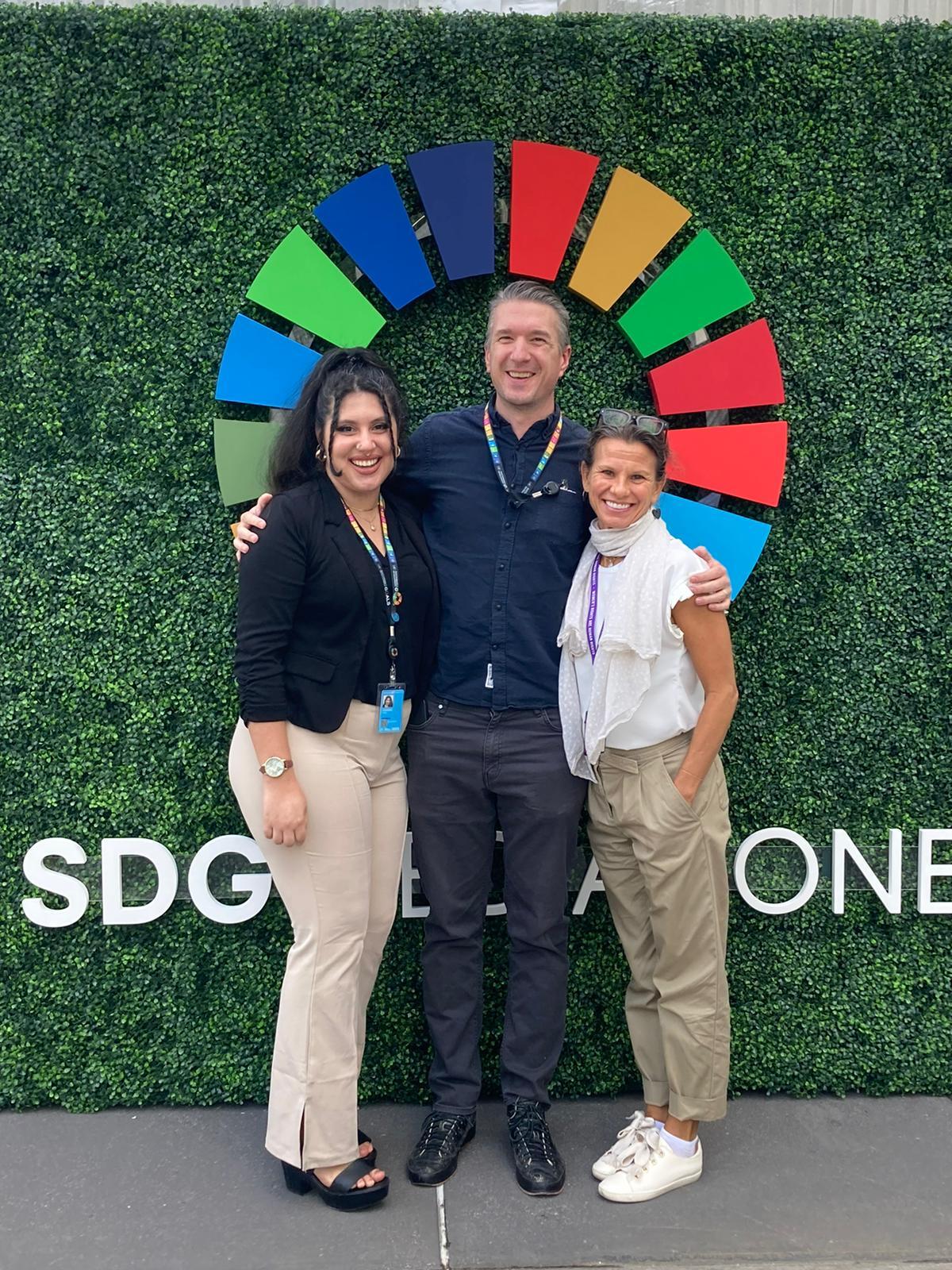 Jennifer Krottinger (right) stands with a man and a woman in front of a faux greenery background with the UN's SDG logo