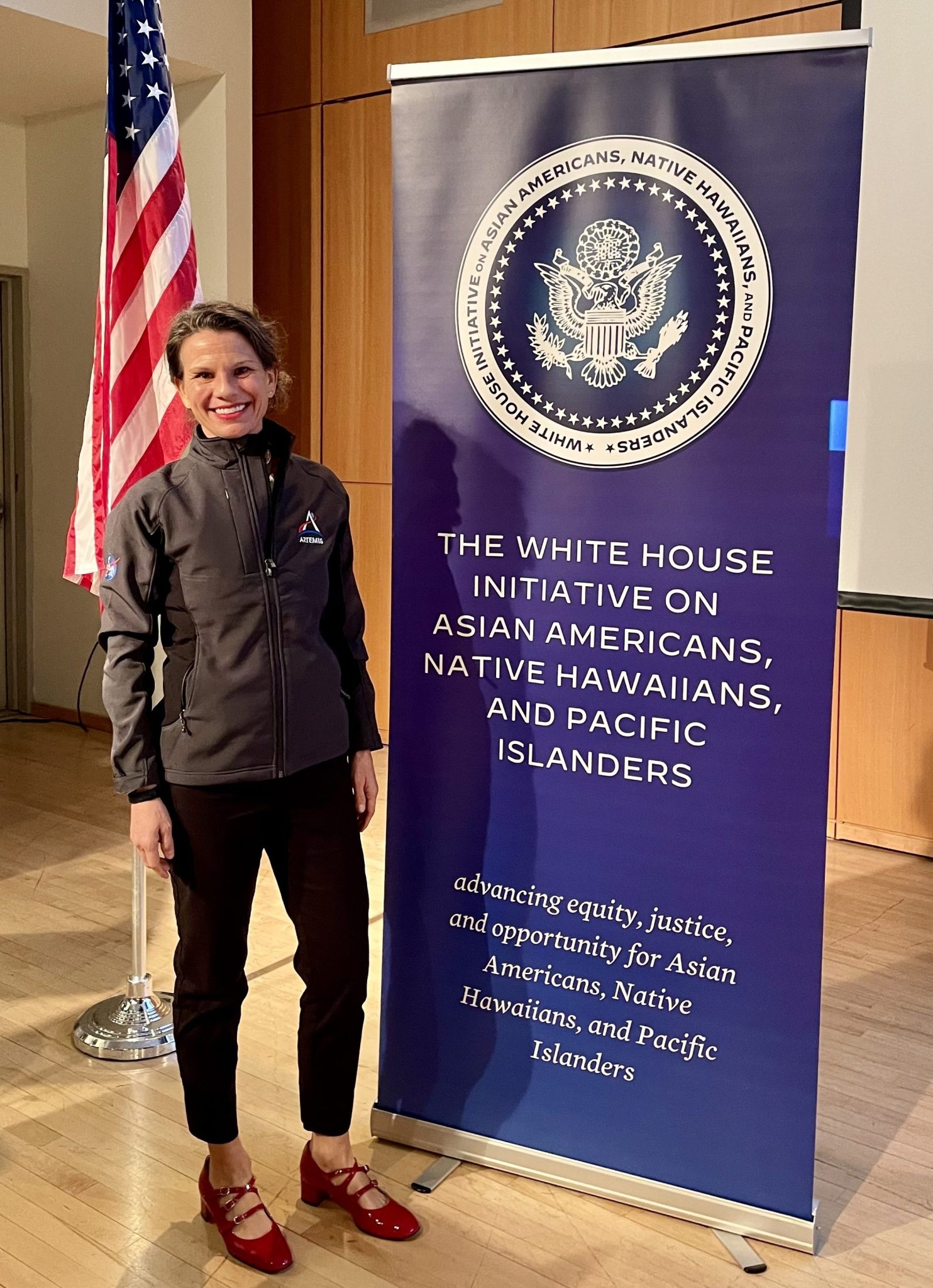 Jennifer Krottinger wears a grey Artemis jacket with black pants and red shoes and smiles next to a sign that says "The White House Initiative on Asian Americans, Native Hawaiians, and Pacific Islanders." An American flag is behind her.