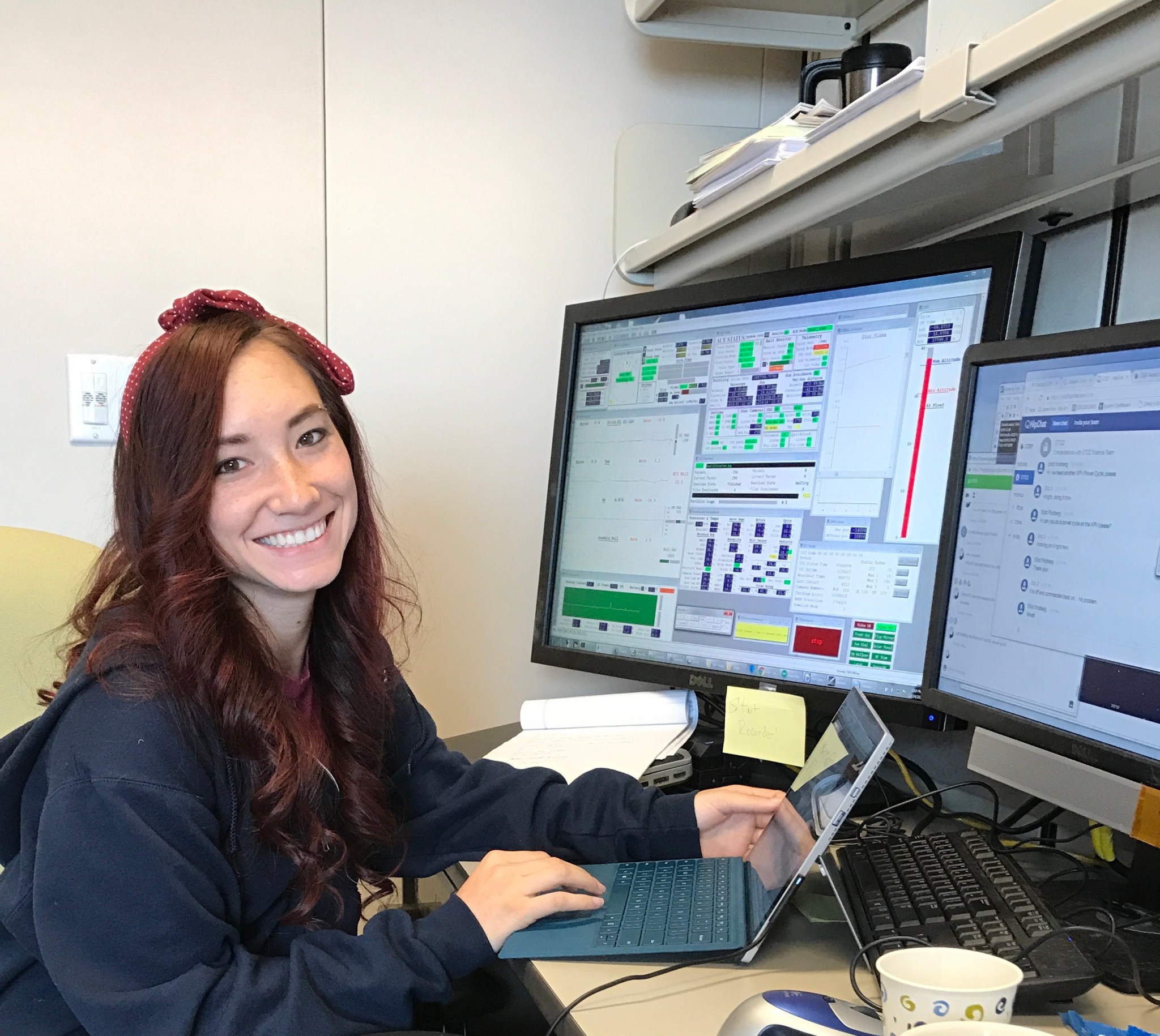 Casey Honniball smiles in front of three computer screens as she works